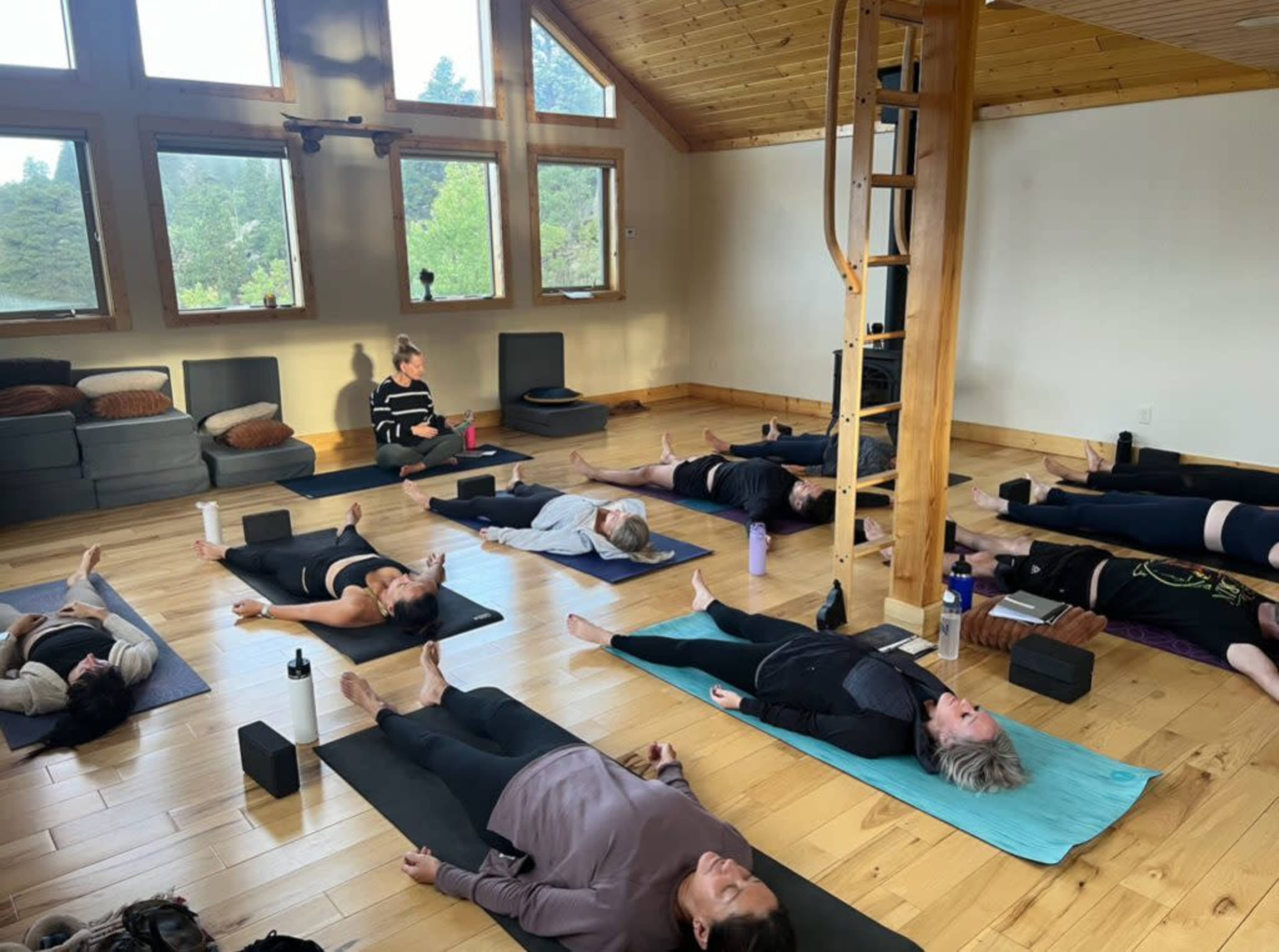 A group of people is practicing yoga in a spacious studio with large windows, surrounded by natural light and wooden accents.