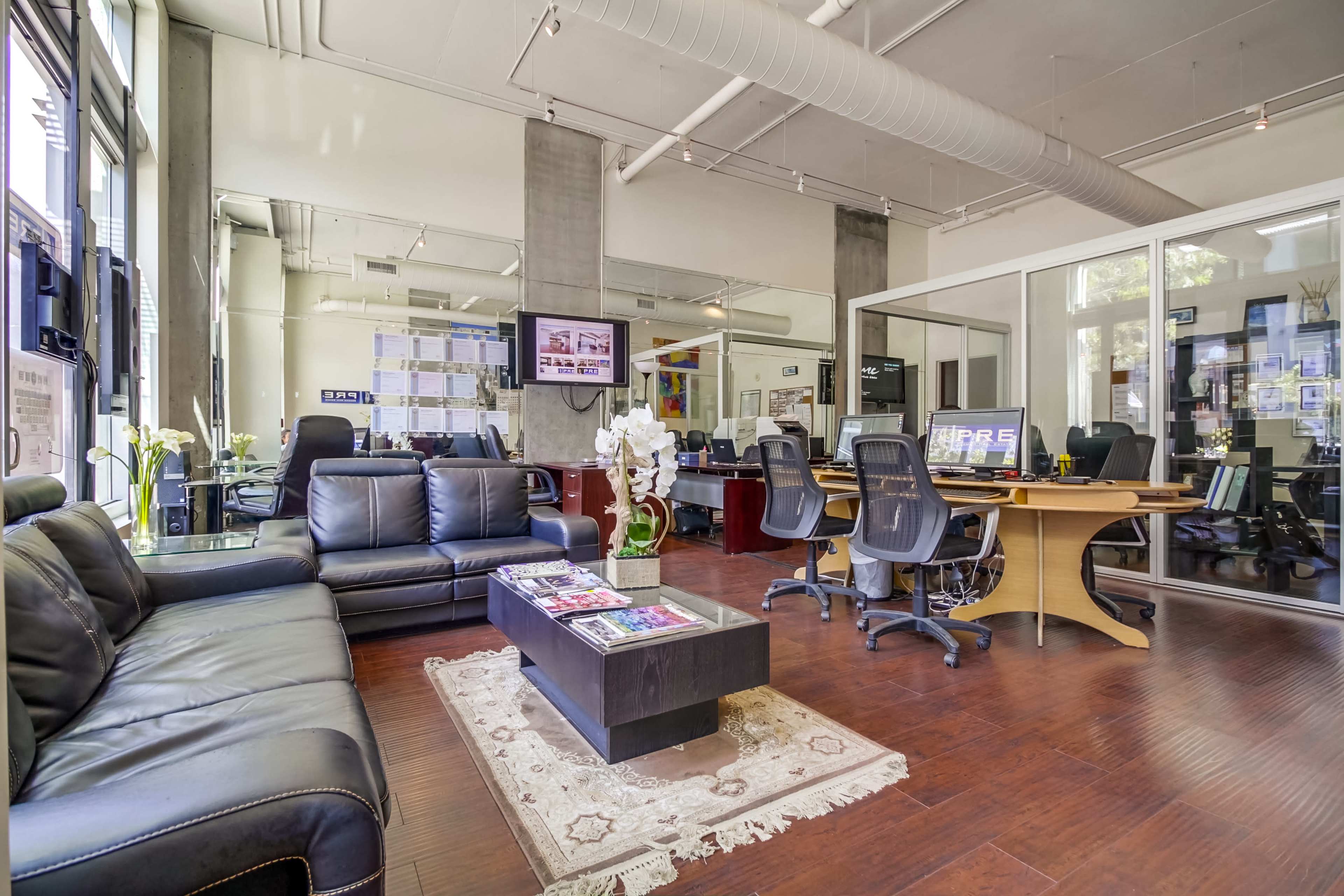 A modern office space features a black leather sofa, a coffee table with magazines, and multiple desks arranged in a bright, open layout.