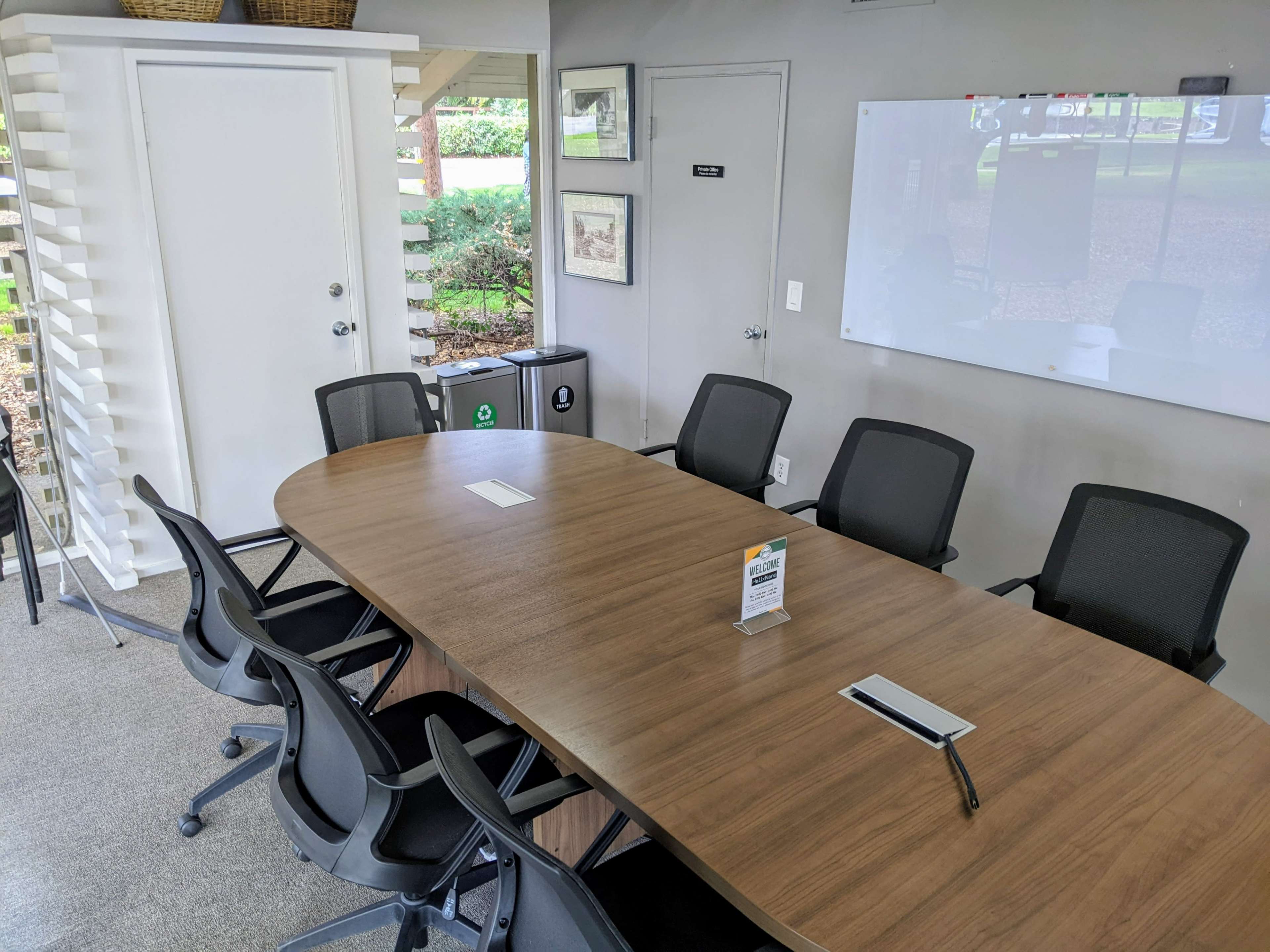 A conference room features a large wooden table surrounded by black office chairs, with a whiteboard and a door leading to a landscaped area visible in the background.