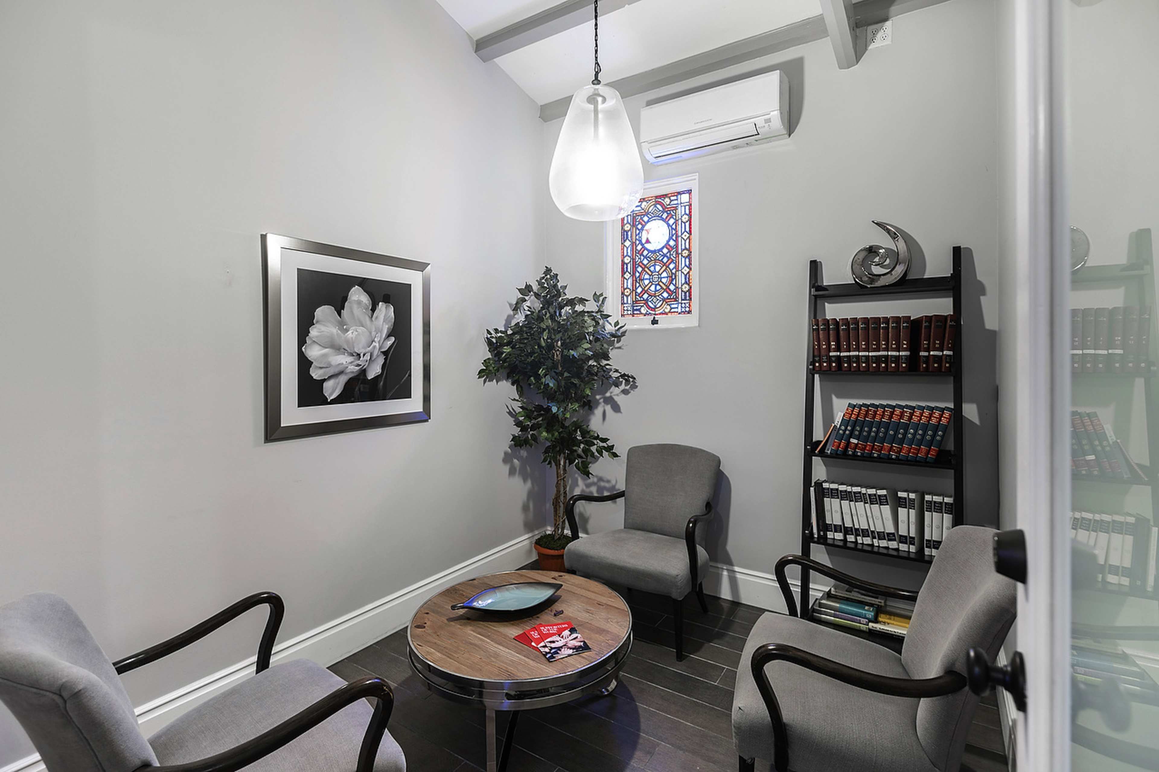 A small room features two armchairs, a coffee table, a bookshelf filled with books, and a decorative plant, with a stained glass window adding color to the gray walls.