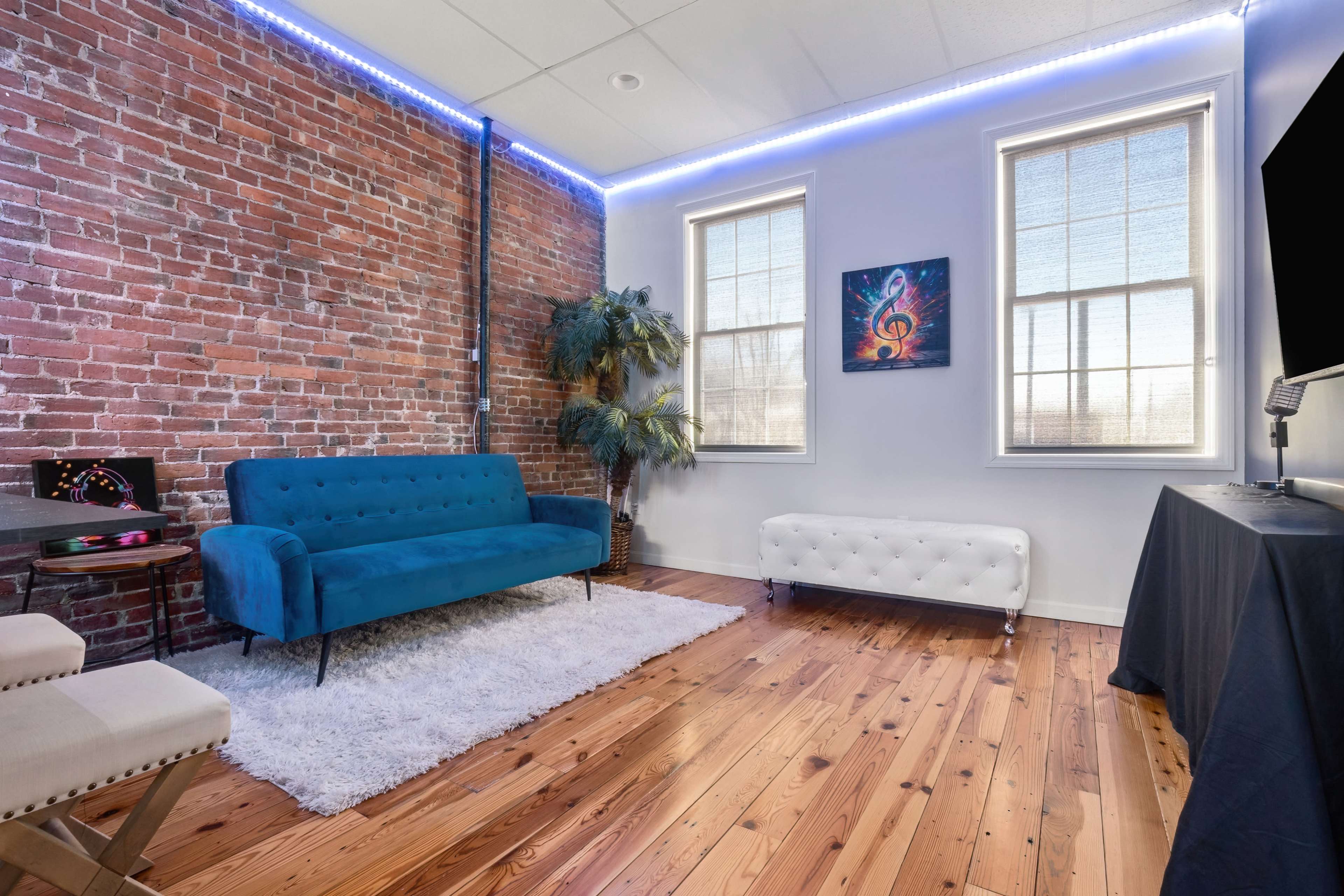 The image shows a room with exposed brick walls, hardwood flooring, a blue couch, a white bench, and two windows letting in natural light.