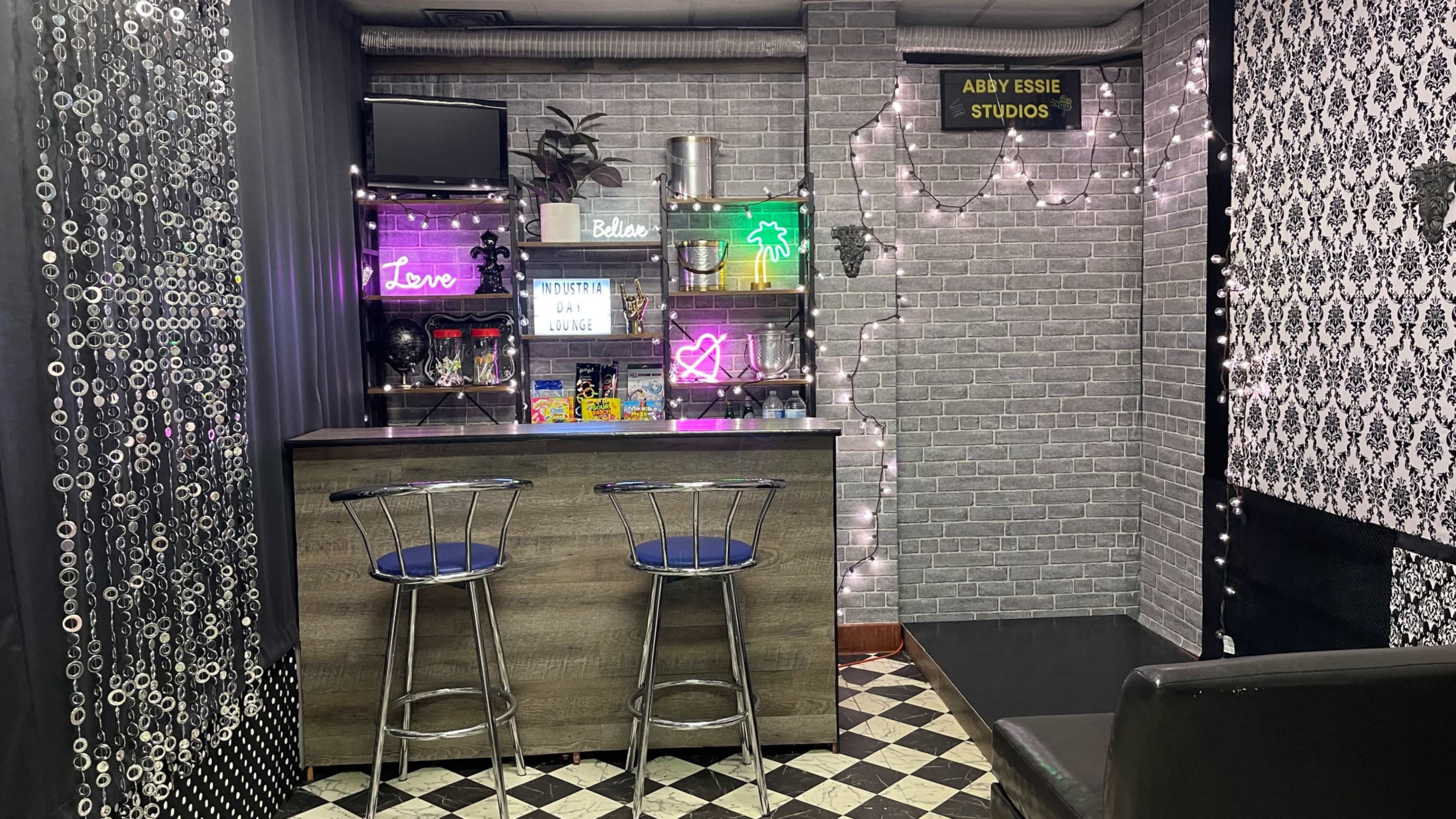 The image shows a stylish studio space with a bar area featuring two stools, neon signs, and a decorative backdrop.