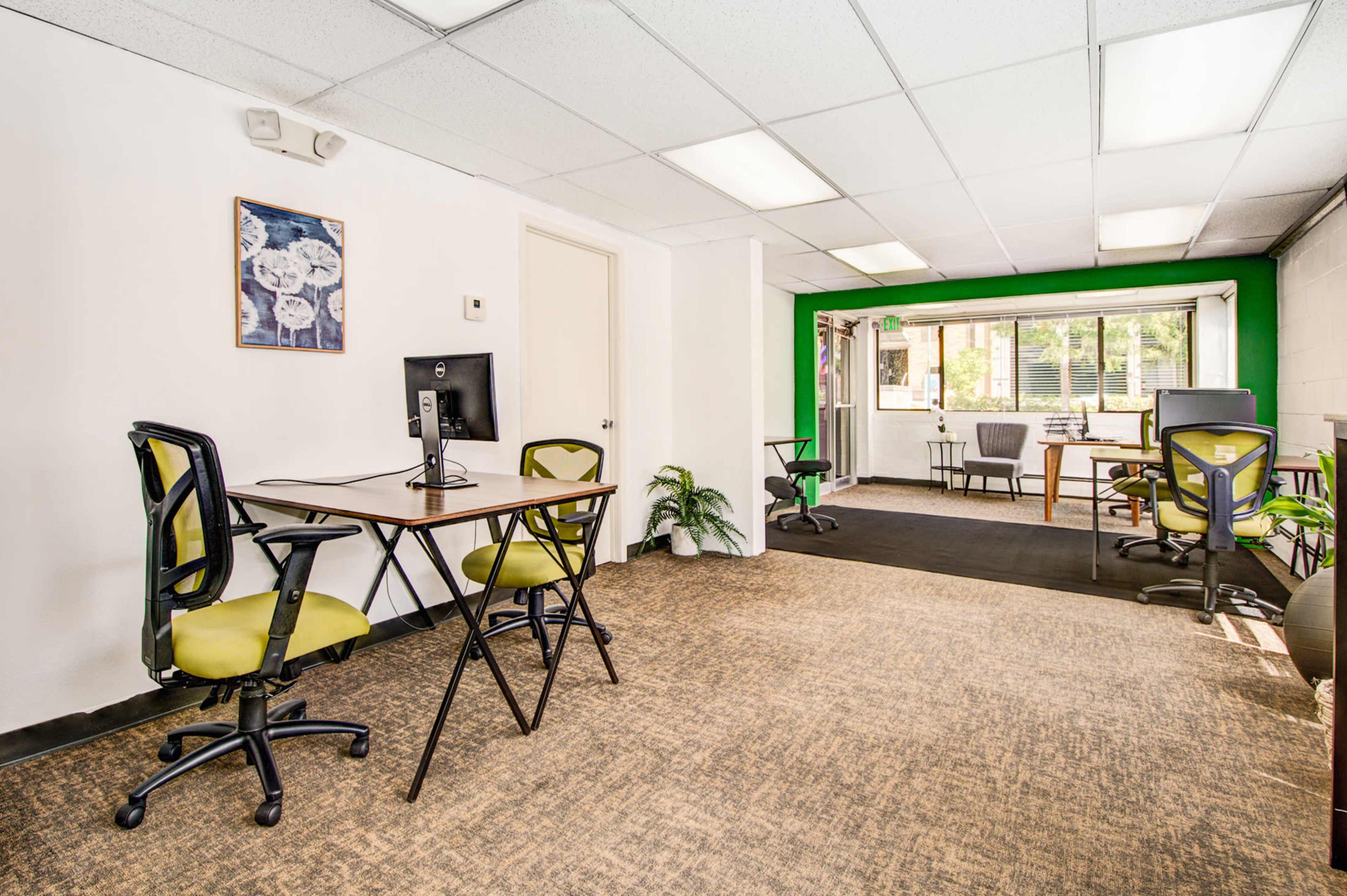 A modern office space with two workstations, featuring black chairs and a desk with a computer, alongside a meeting area in the background.