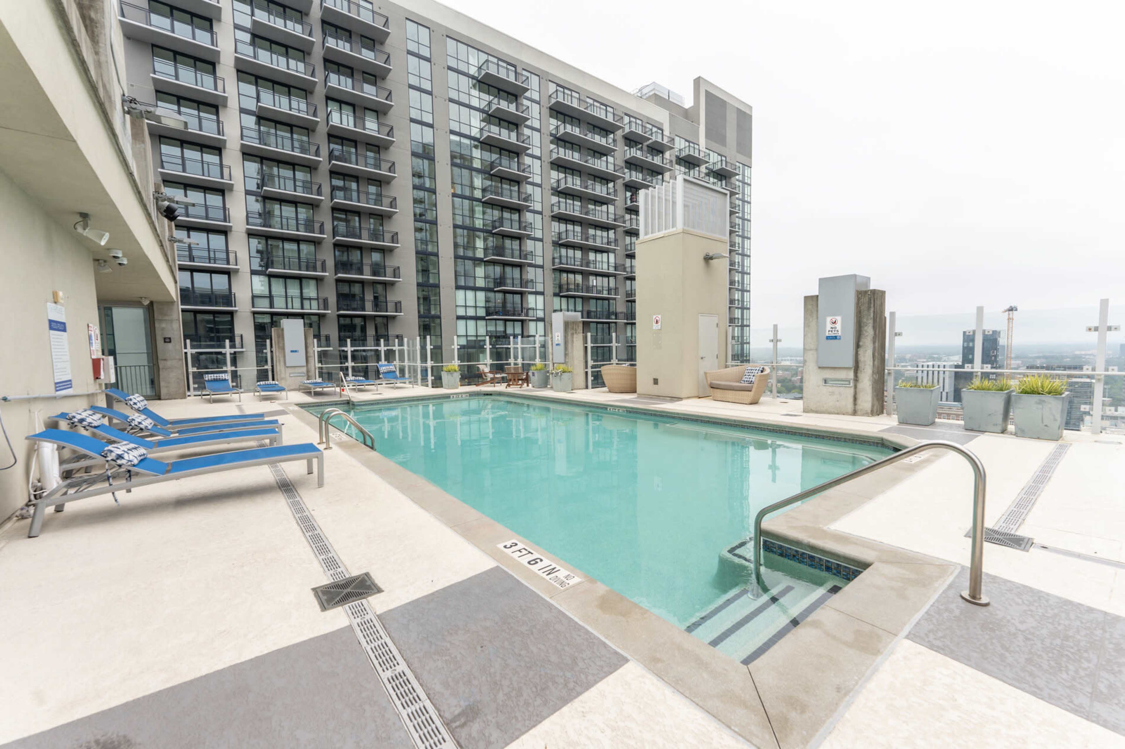 The image shows a modern rooftop pool area surrounded by high-rise buildings with lounge chairs on one side.