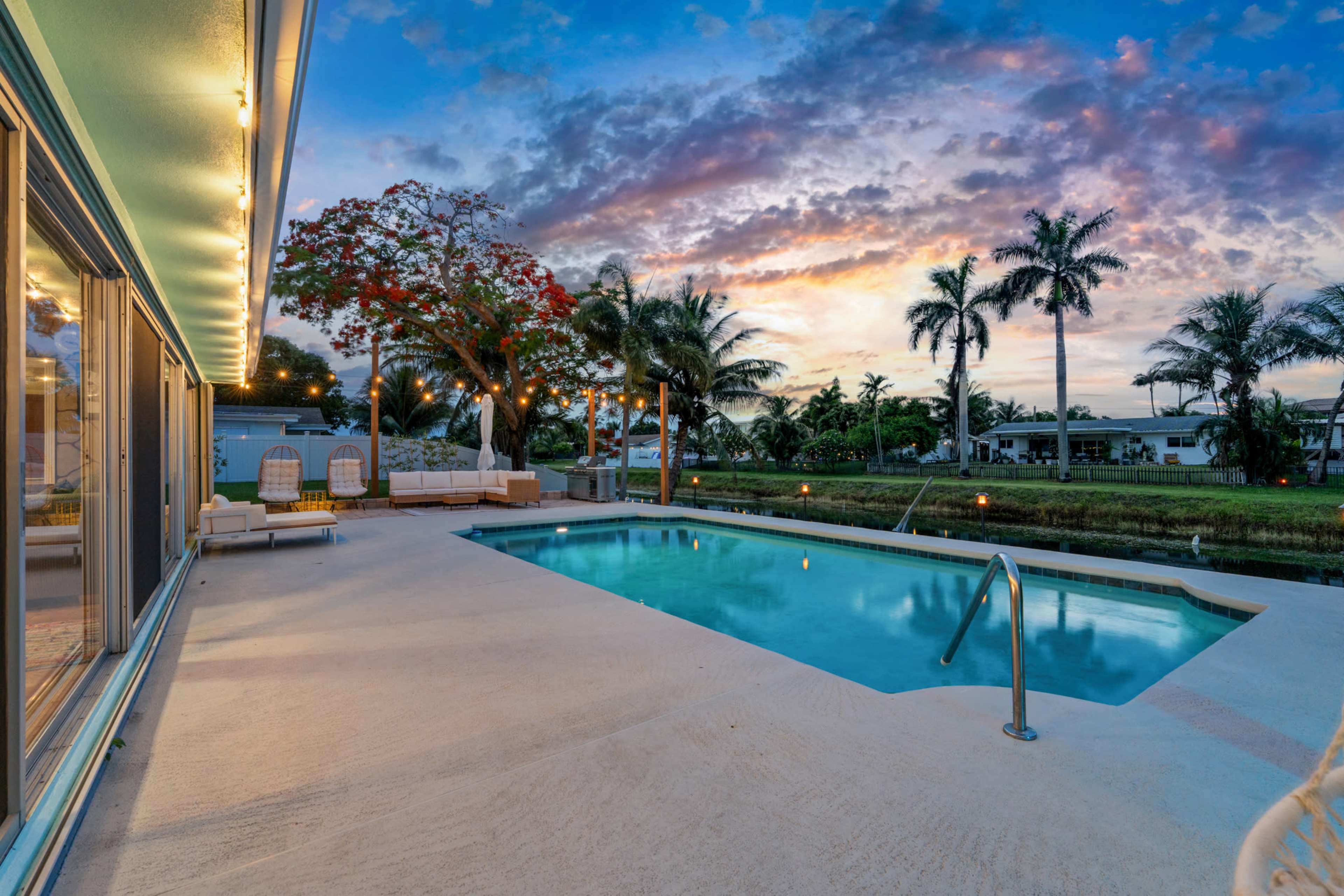 The image shows a swimming pool surrounded by a patio with lounge chairs, set against a colorful sunset sky and palm trees.