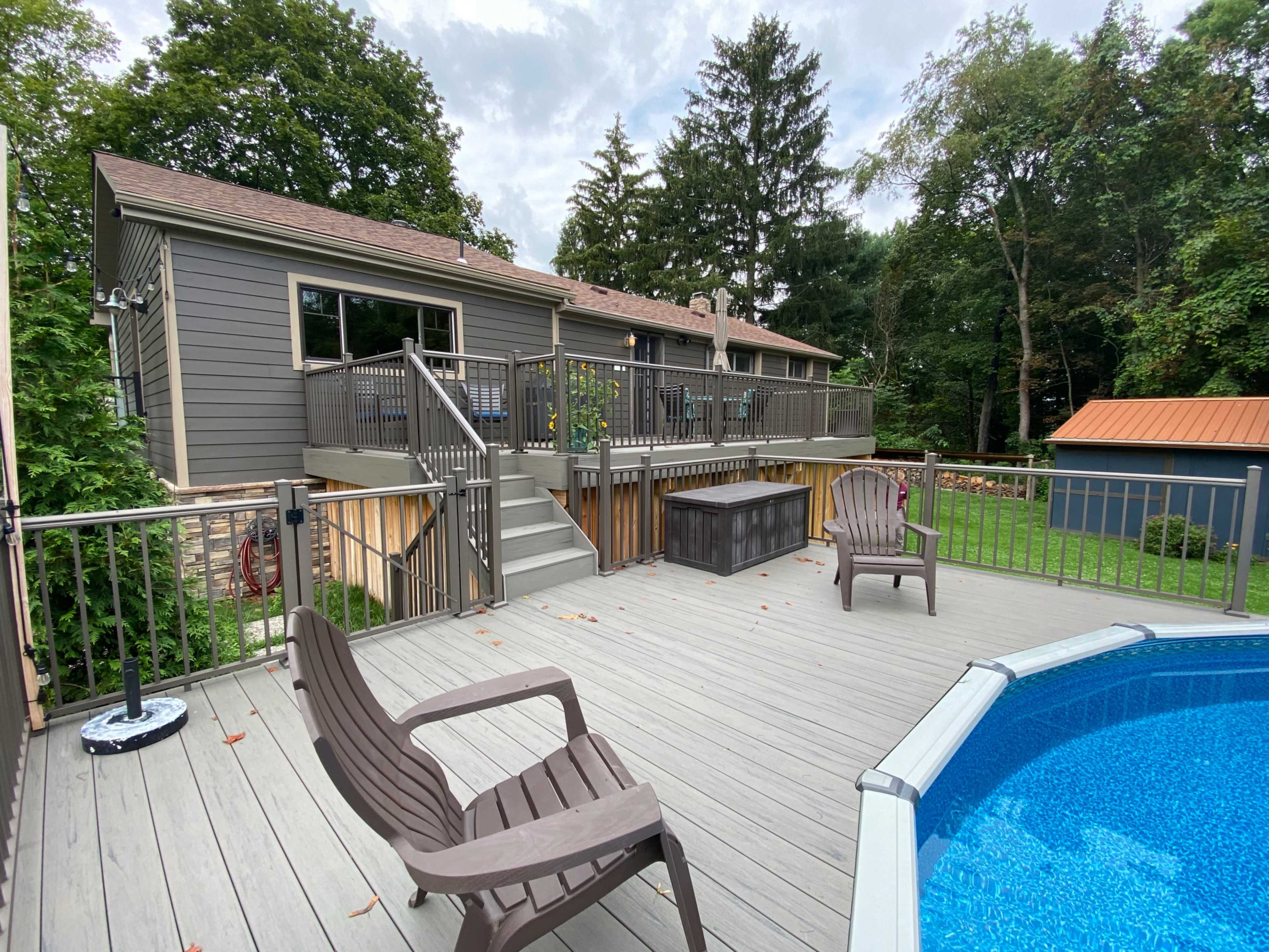 A wooden deck with two chairs overlooks a blue above-ground pool and a house surrounded by trees.
