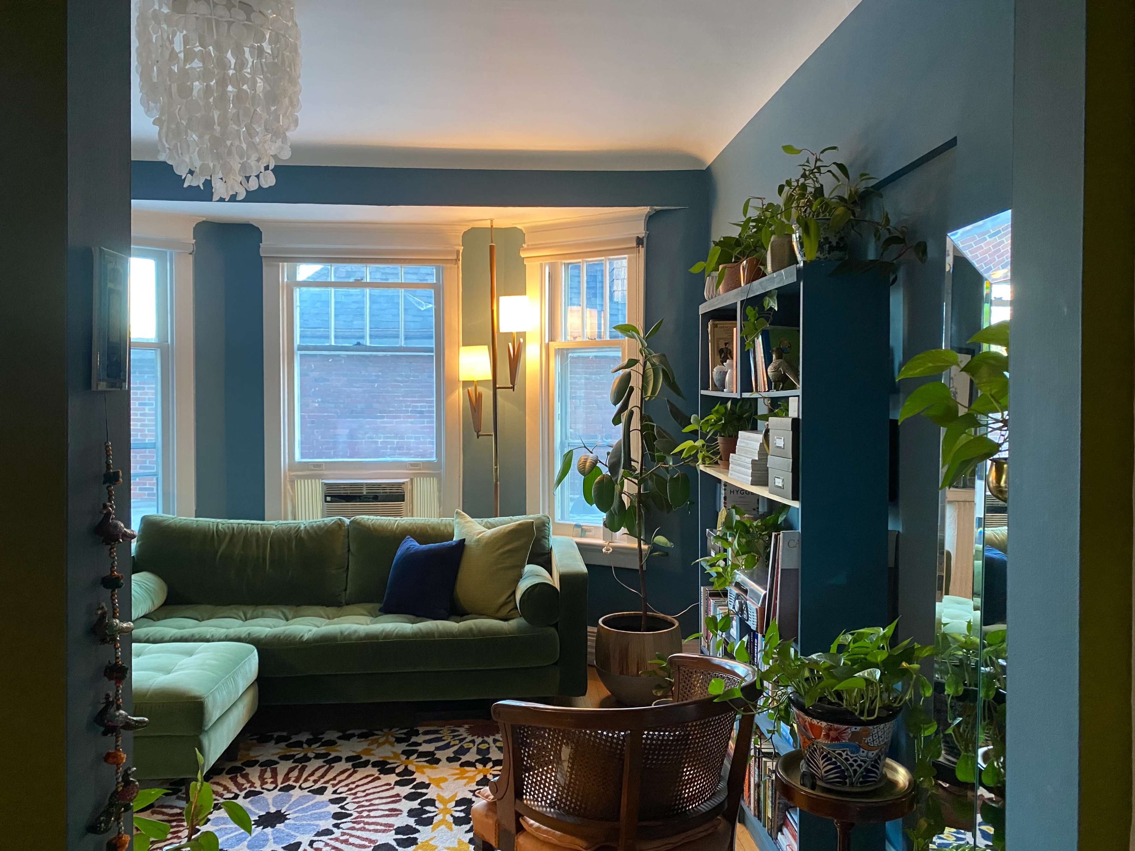 A cozy living room with a green sofa, a wooden chair, plants on shelves, and large windows allowing natural light to enter.