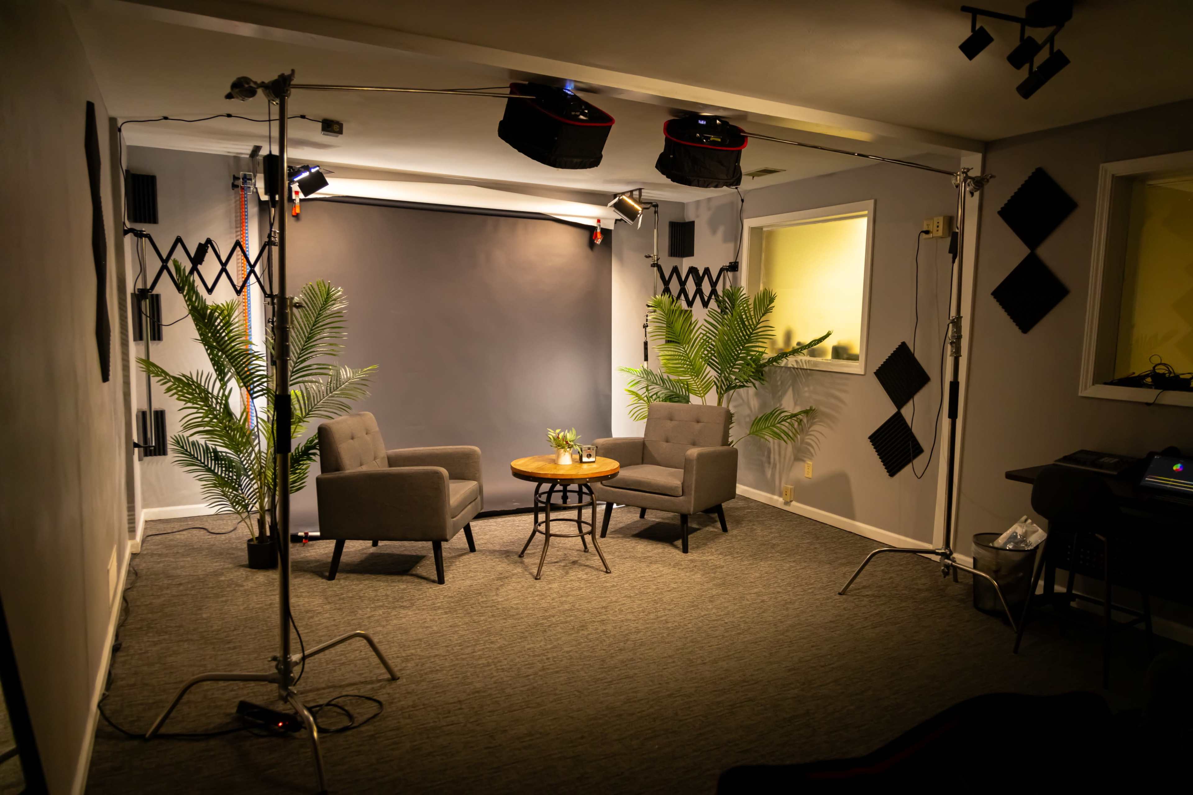 The image shows a small studio setup with two gray chairs facing a round table, surrounded by potted plants and studio lighting equipment.