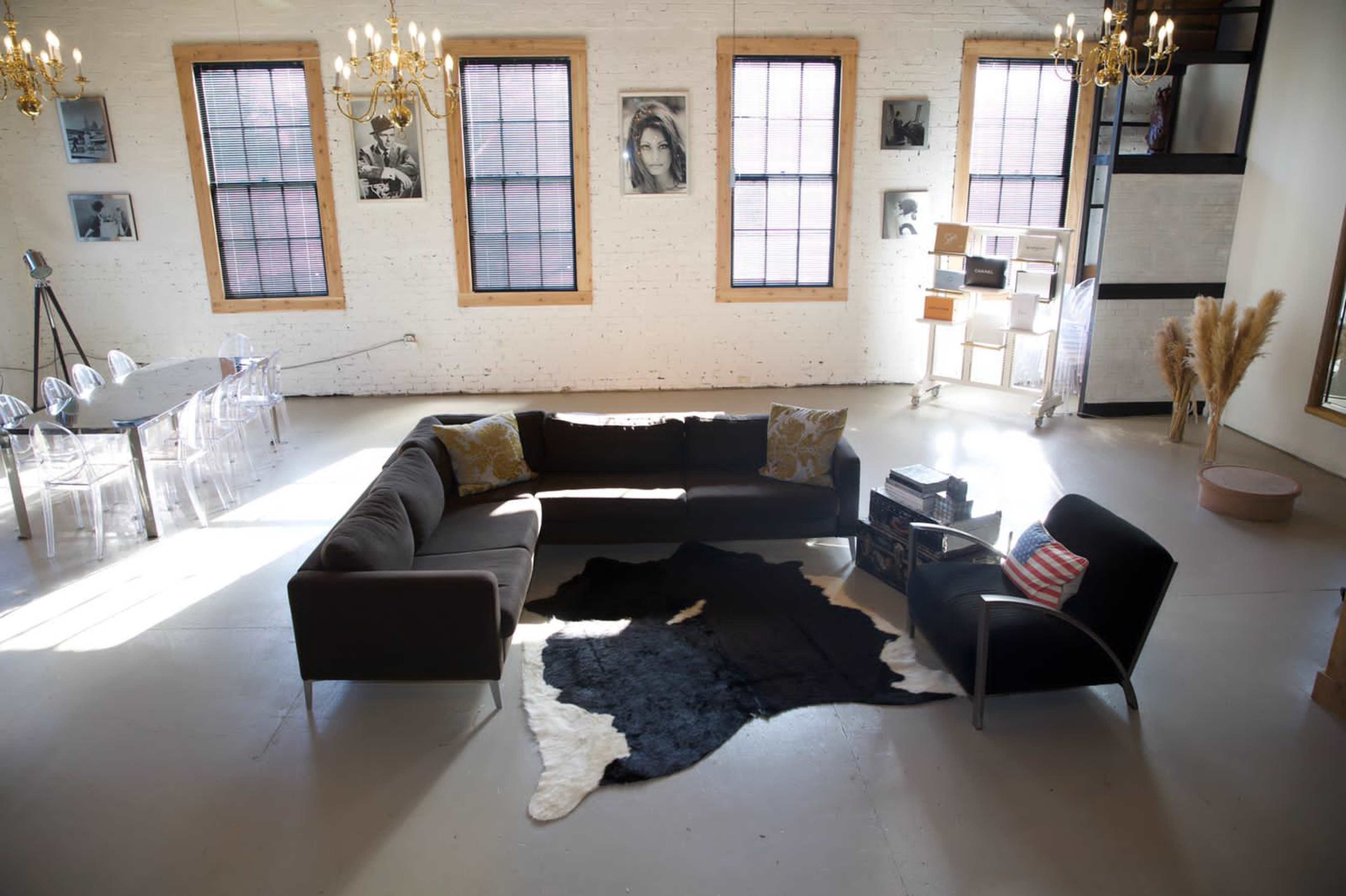 A spacious, well-lit living area featuring a sectional sofa, a coffee table, and a decorative rug, surrounded by large windows and artworks on the walls.