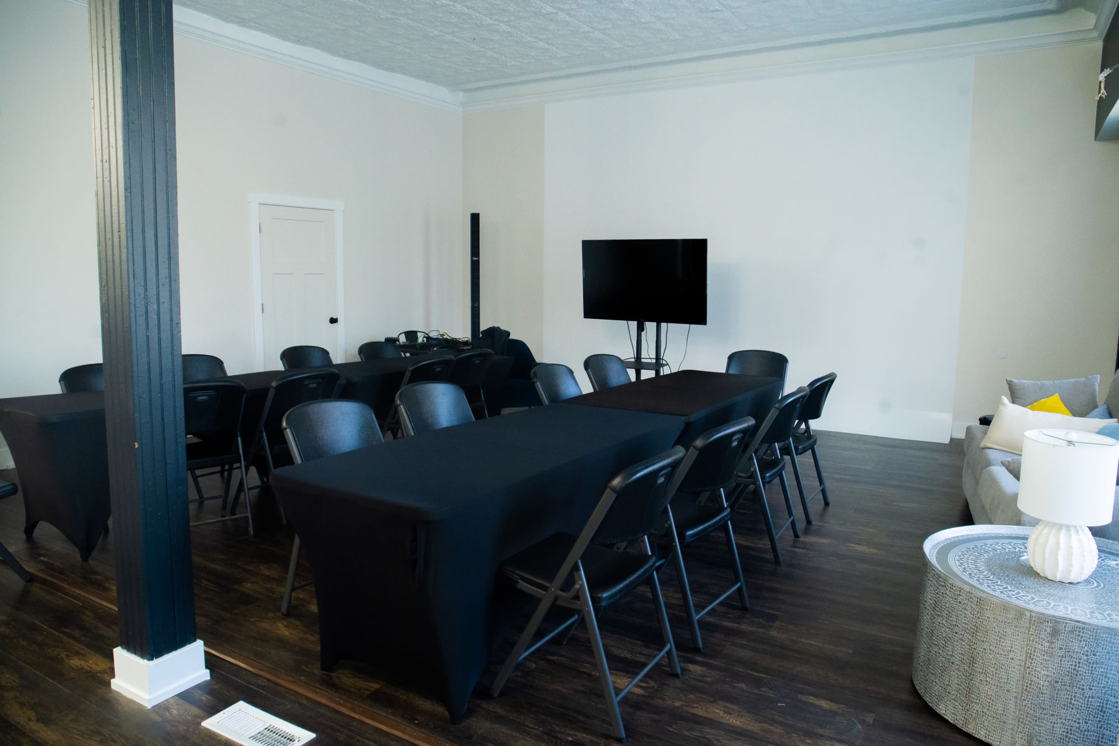 A conference room is set up with several black tables and chairs arranged in a rectangular layout, featuring a television mounted on the wall.