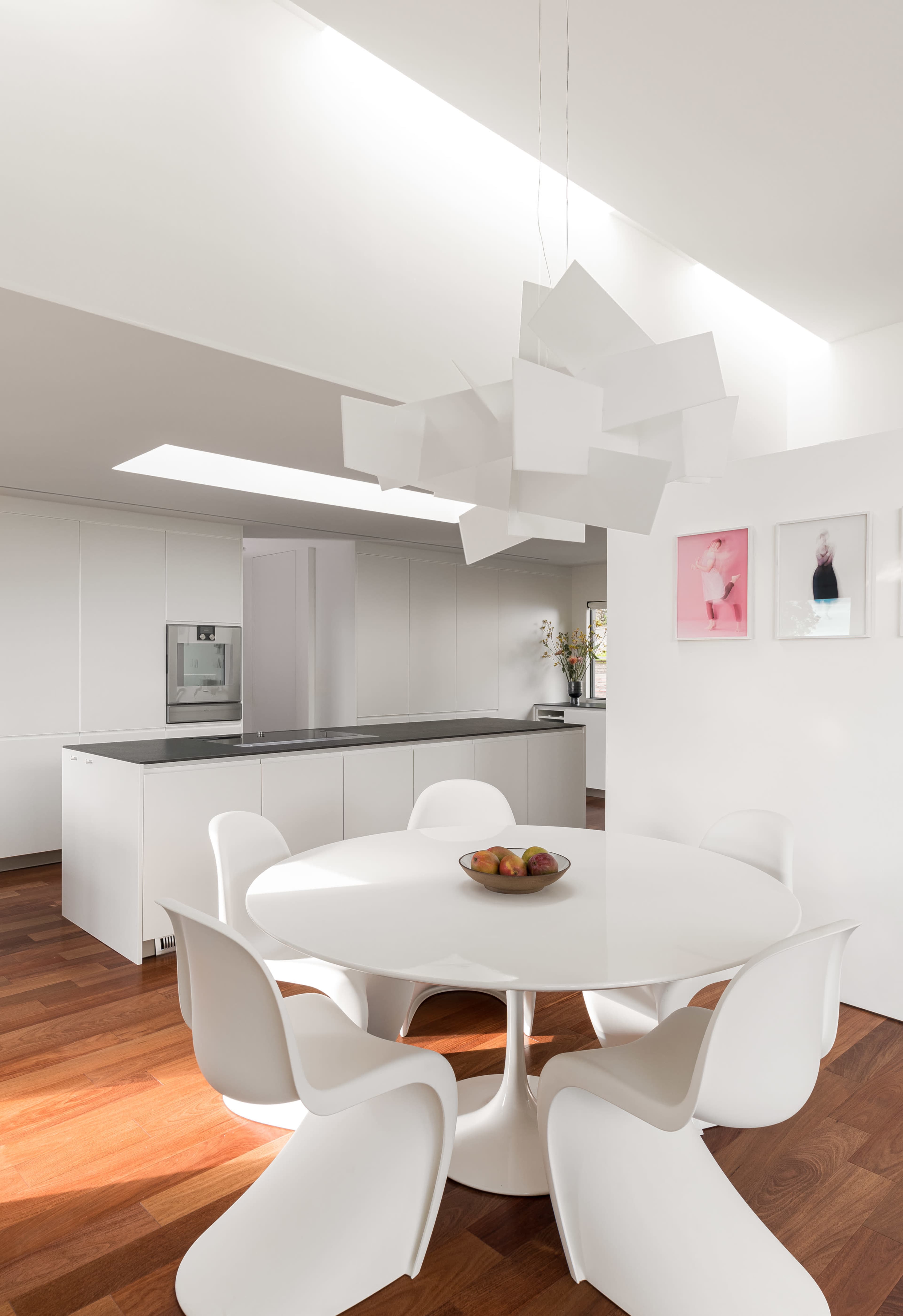 A modern kitchen features a round white table with six chairs, a bowl of fruit at the center, and a unique light fixture overhead.