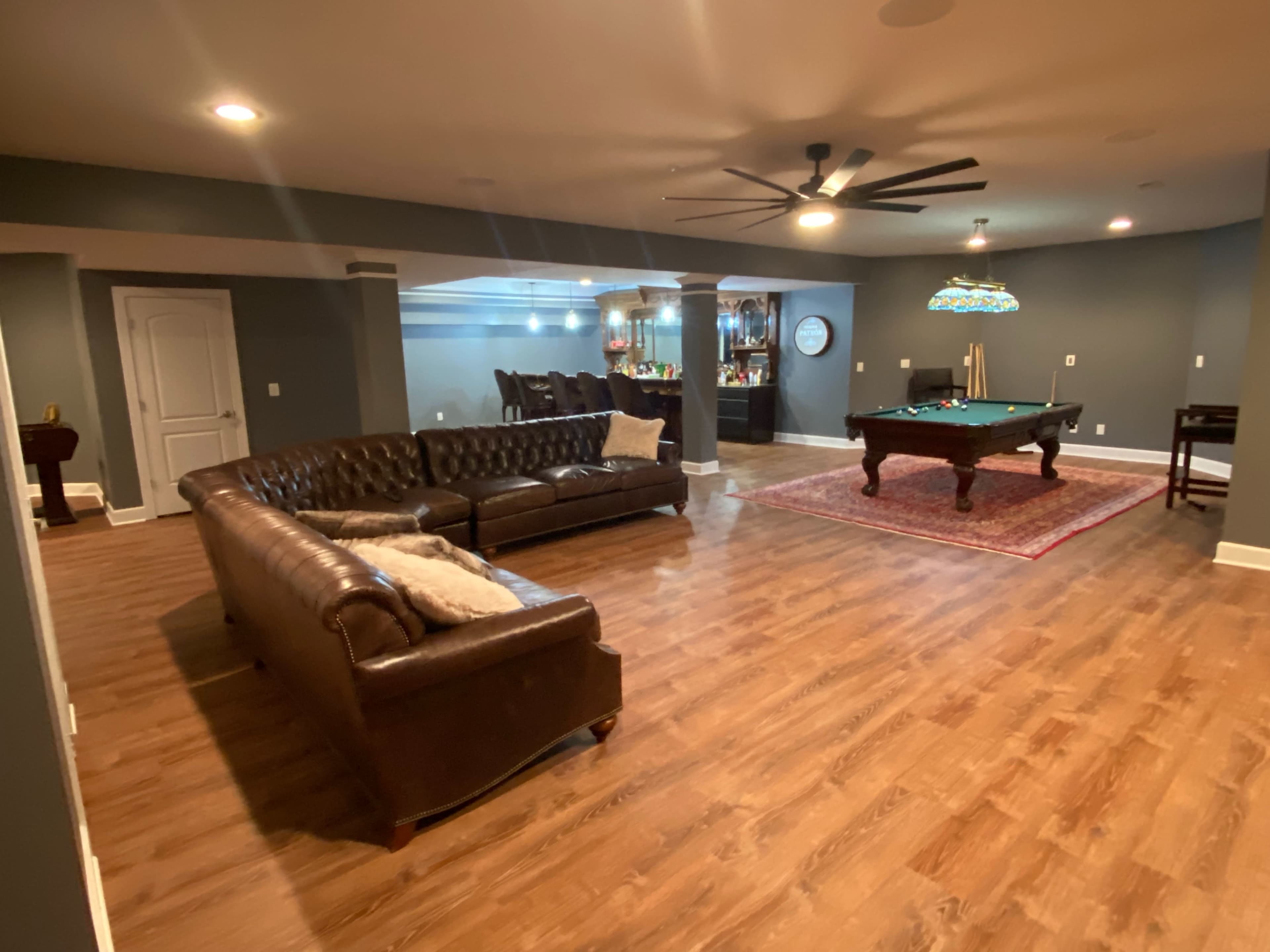 A finished basement with a pool table, a brown leather sectional sofa, and a bar area in the background.