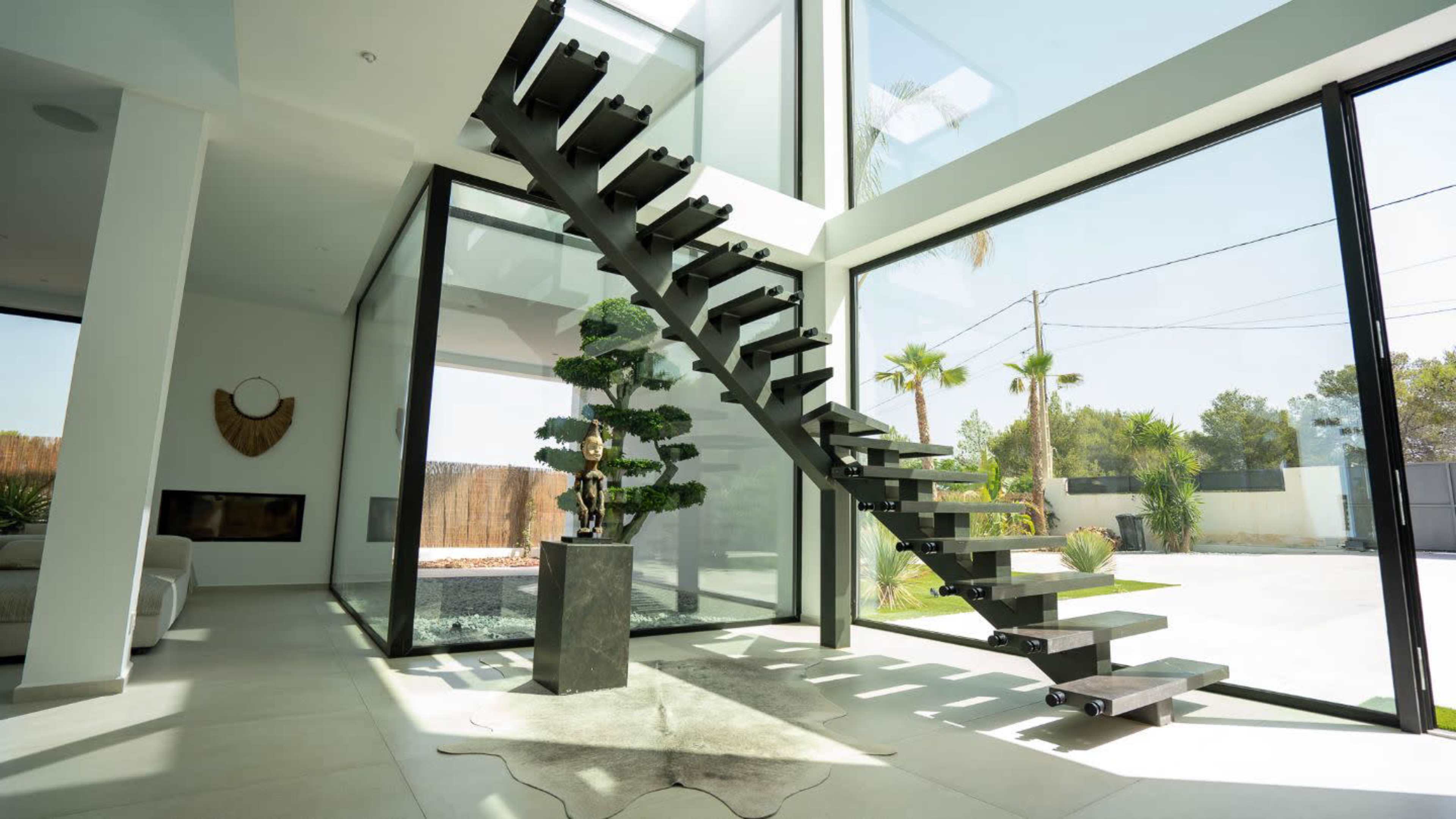 A modern interior featuring a glass-walled space with an open staircase, a planter sculpture, and large windows letting in natural light.