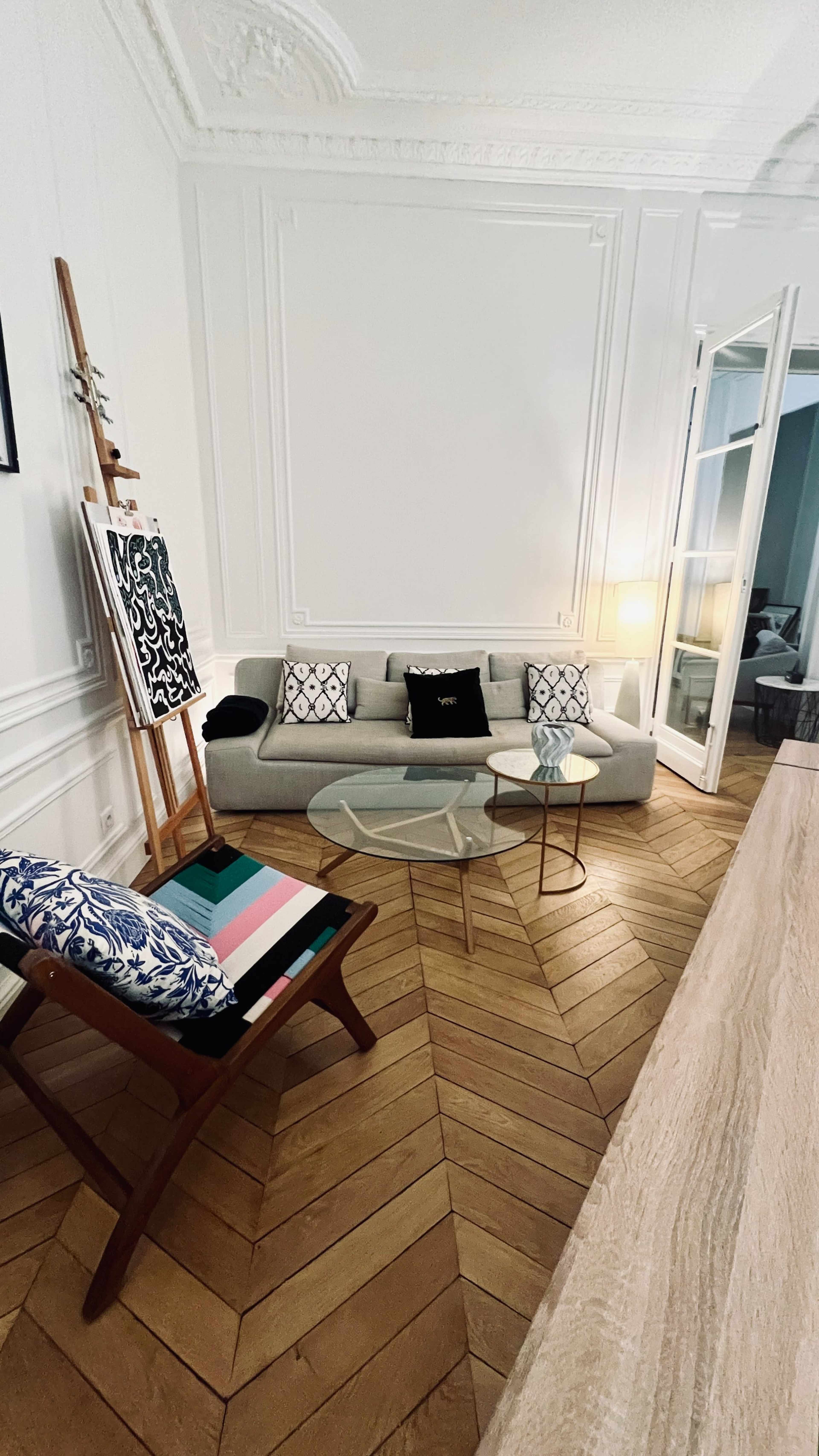The living room features a light gray sofa with patterned cushions, a round glass coffee table, and a wooden chair with colorful seat cushions, all set against a backdrop of herringbone wooden flooring and white walls.