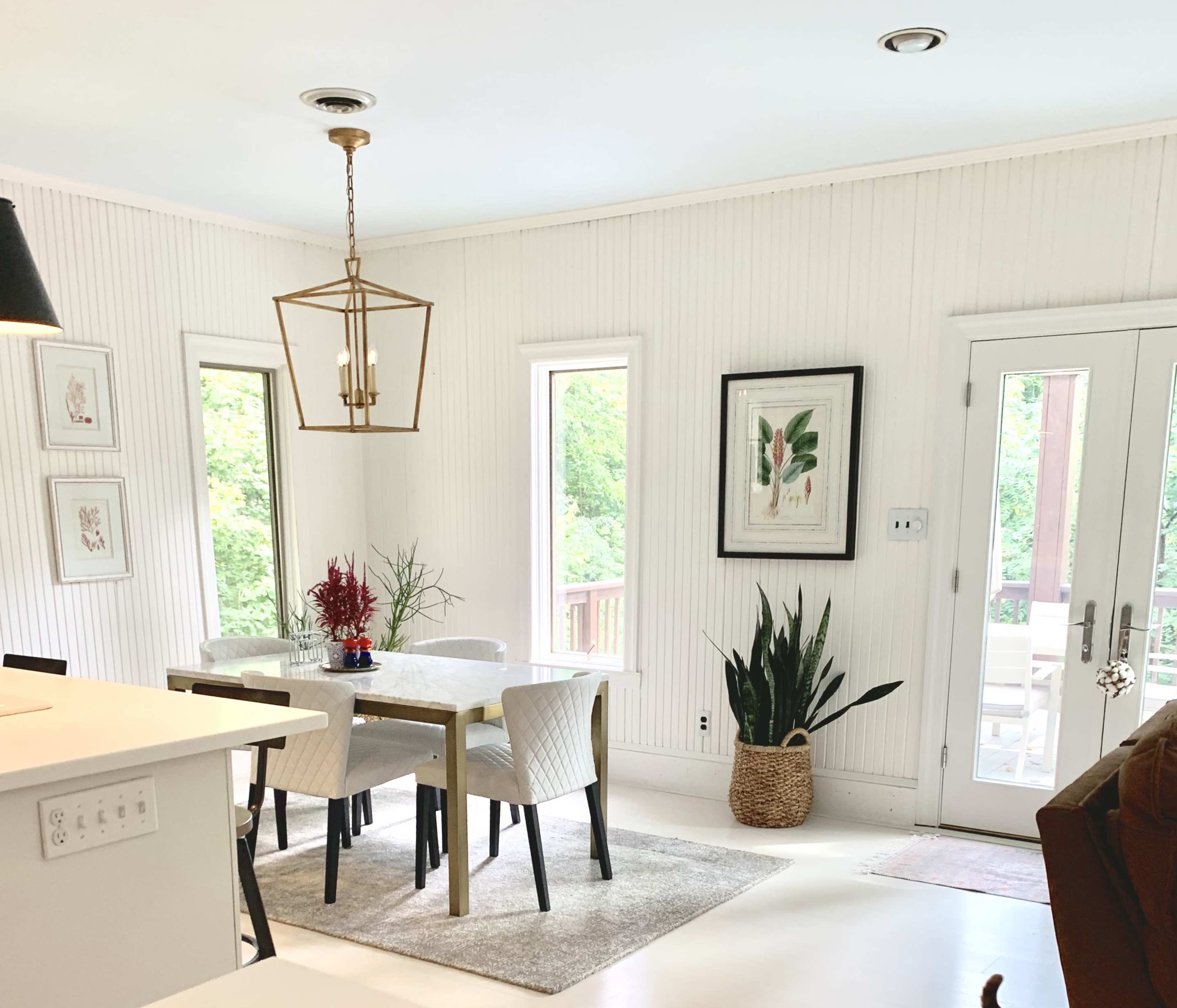 A dining area features a table surrounded by chairs, with large windows and plants, overlooking a wooden deck.