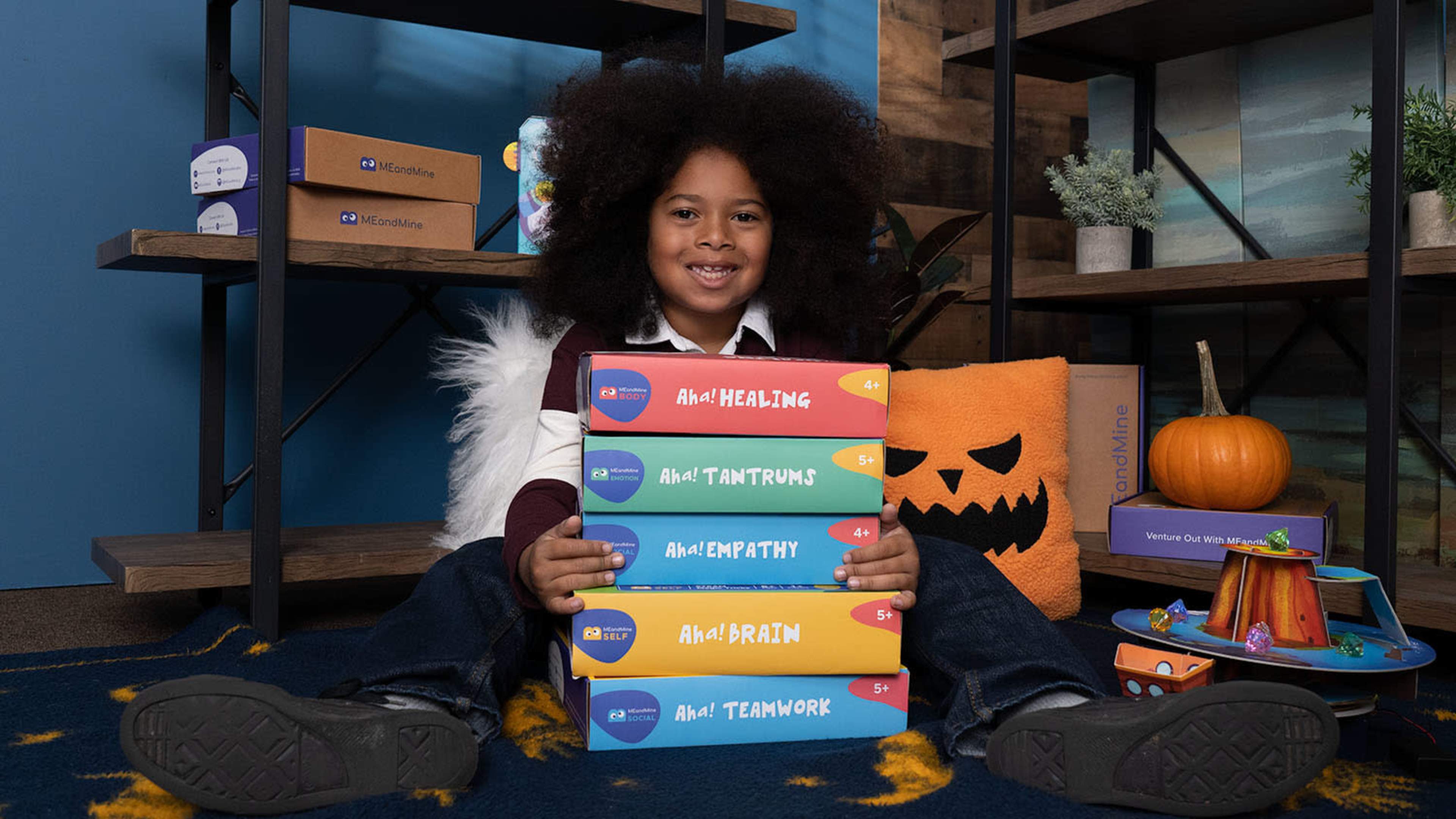 A child with curly hair sits on a rug, holding a stack of colorful books labeled with various titles, surrounded by decorative items including a pumpkin and a plush cushion.