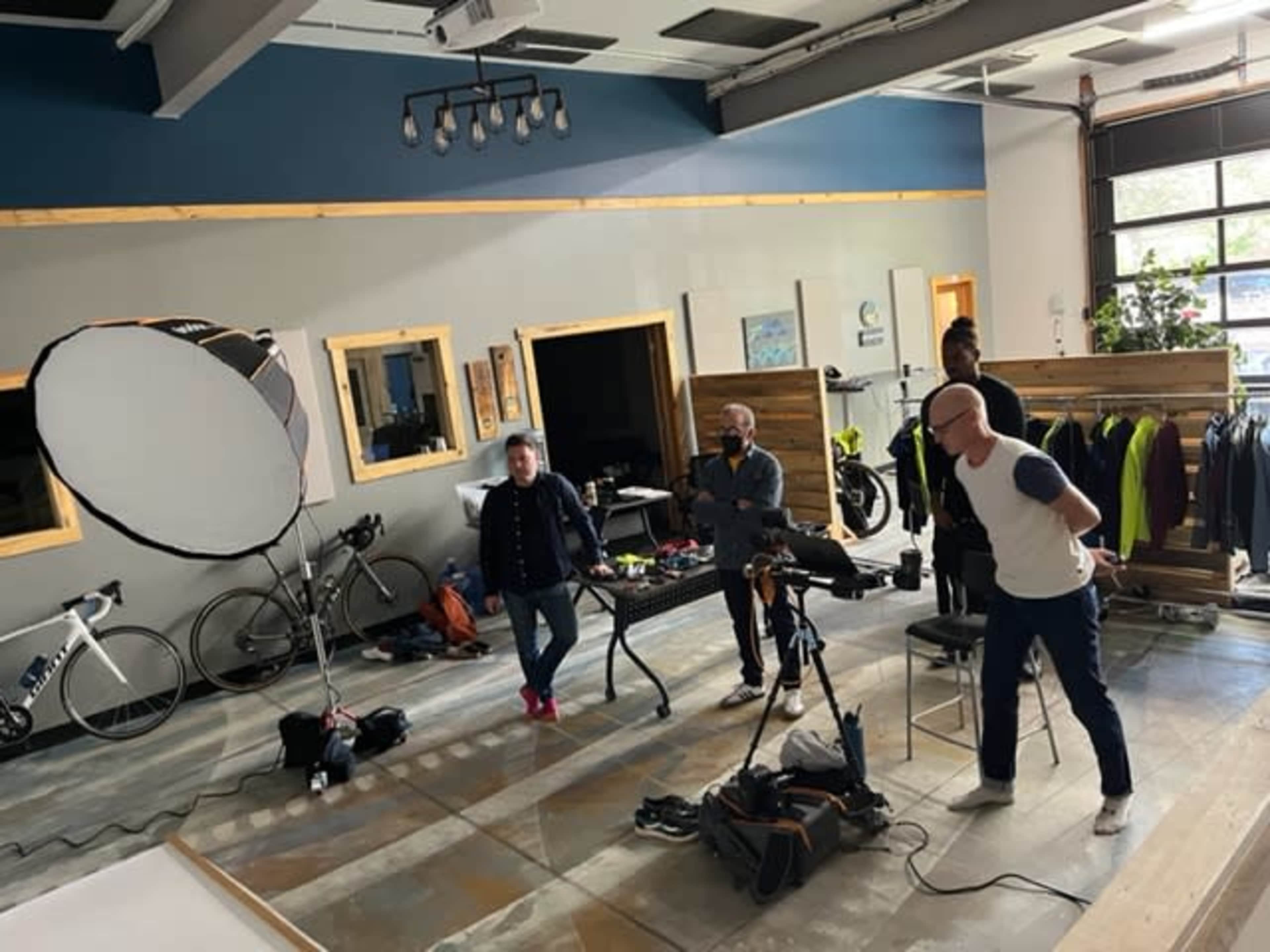 A group of four people is gathered in a studio, with a professional lighting setup and various cycling equipment in the background.