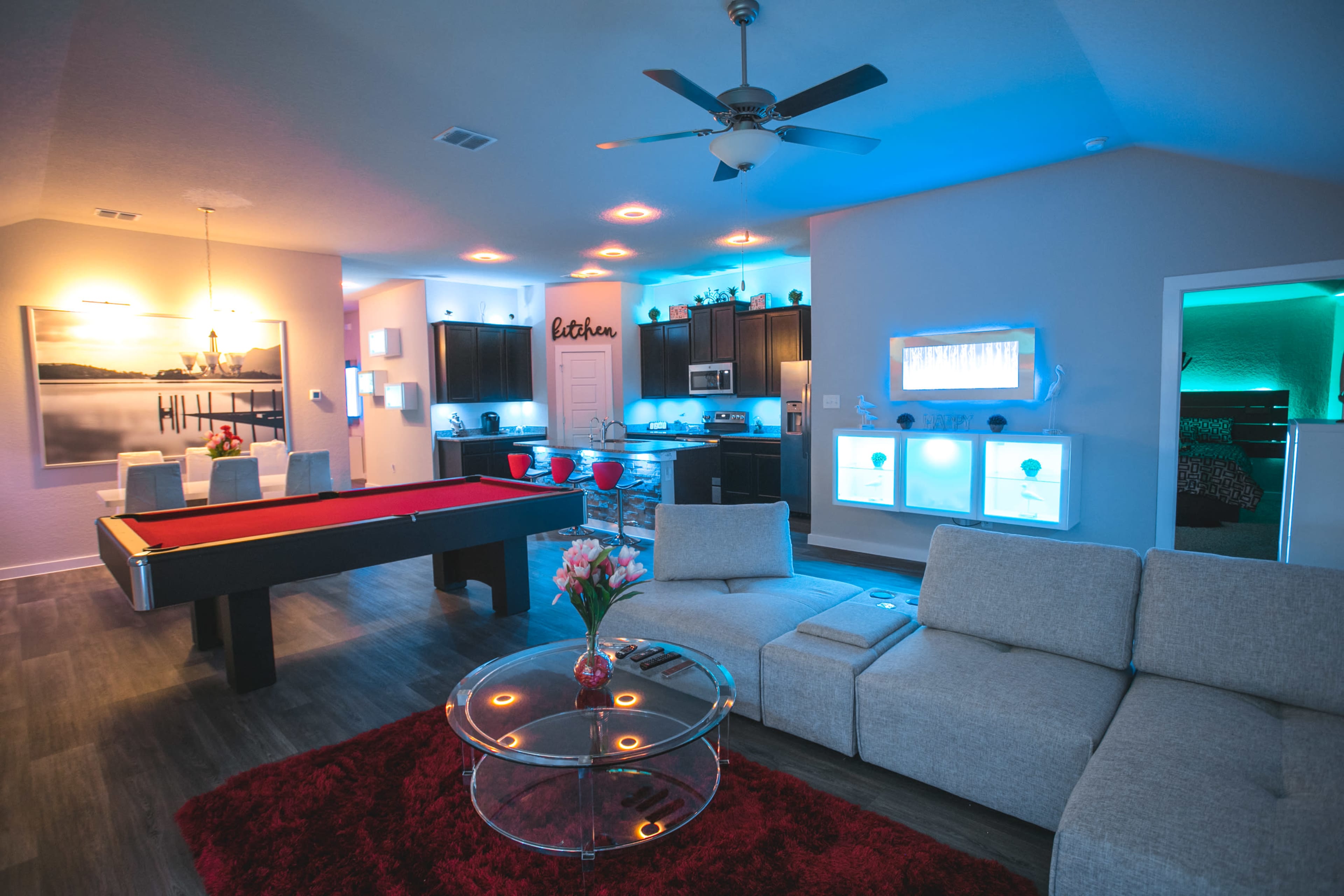 The image shows a modern living room with a pool table, a sectional sofa, and a kitchen area illuminated with blue lighting.