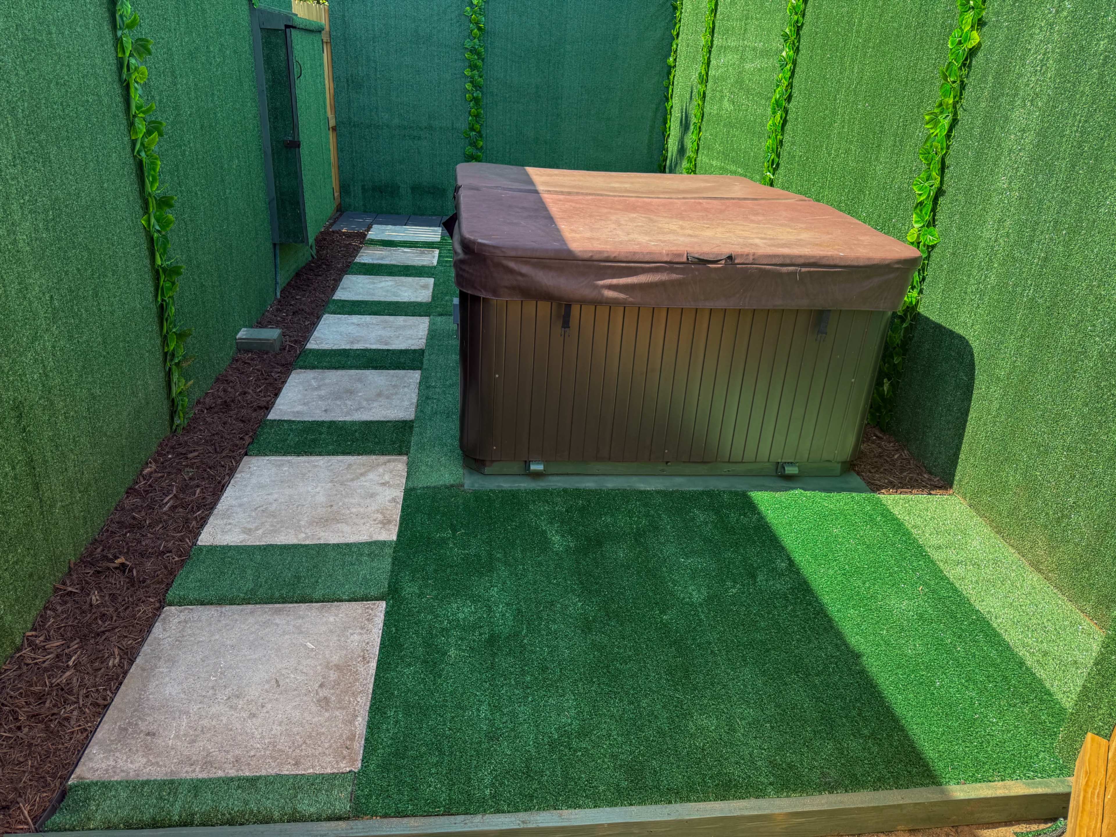 The image shows a fenced outdoor area with green artificial turf, stone pathway, and a hot tub covered with a brown lid.