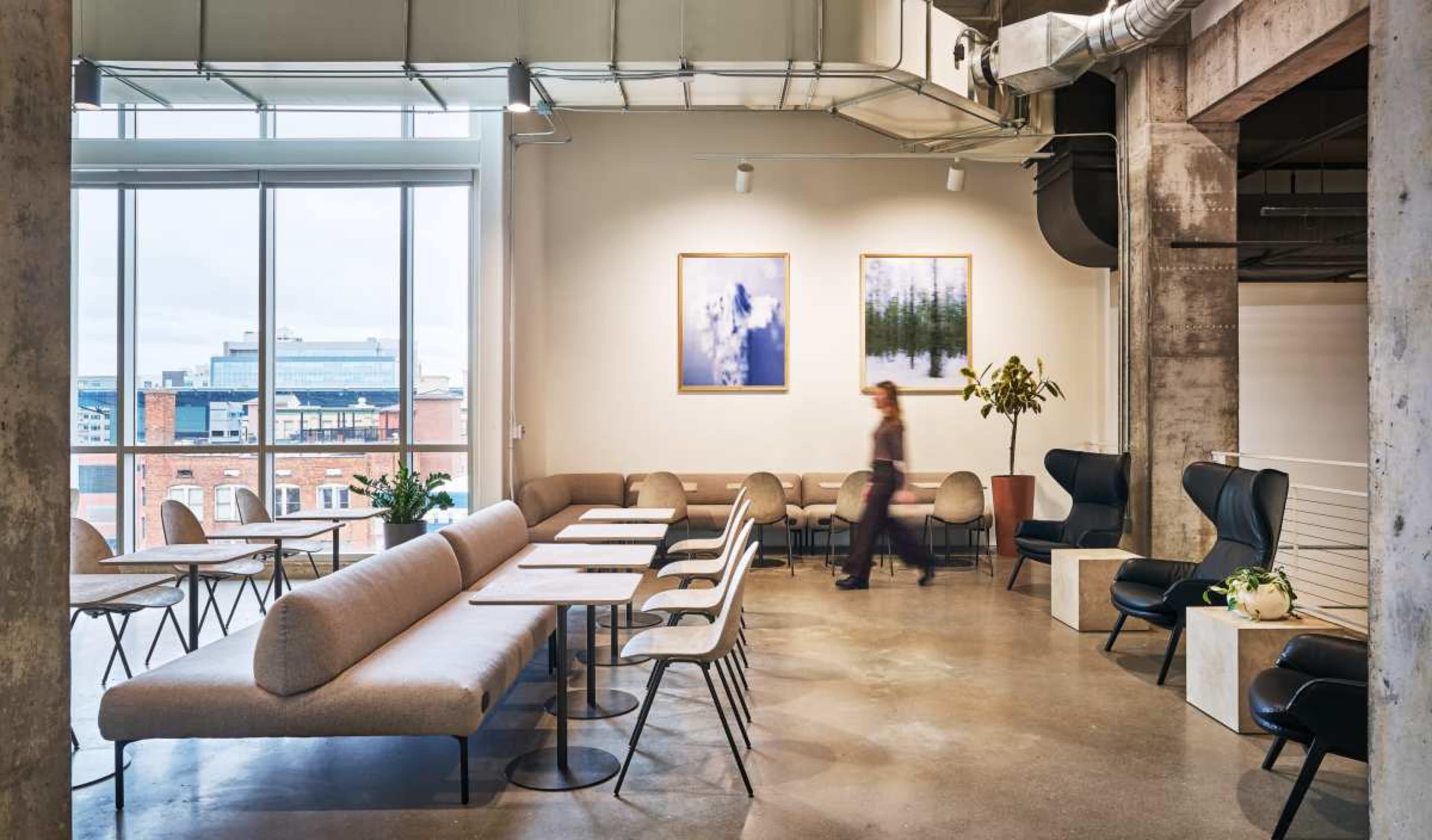 The image depicts a modern lounge area with light-colored seating and tables, large windows, and artwork on the walls.