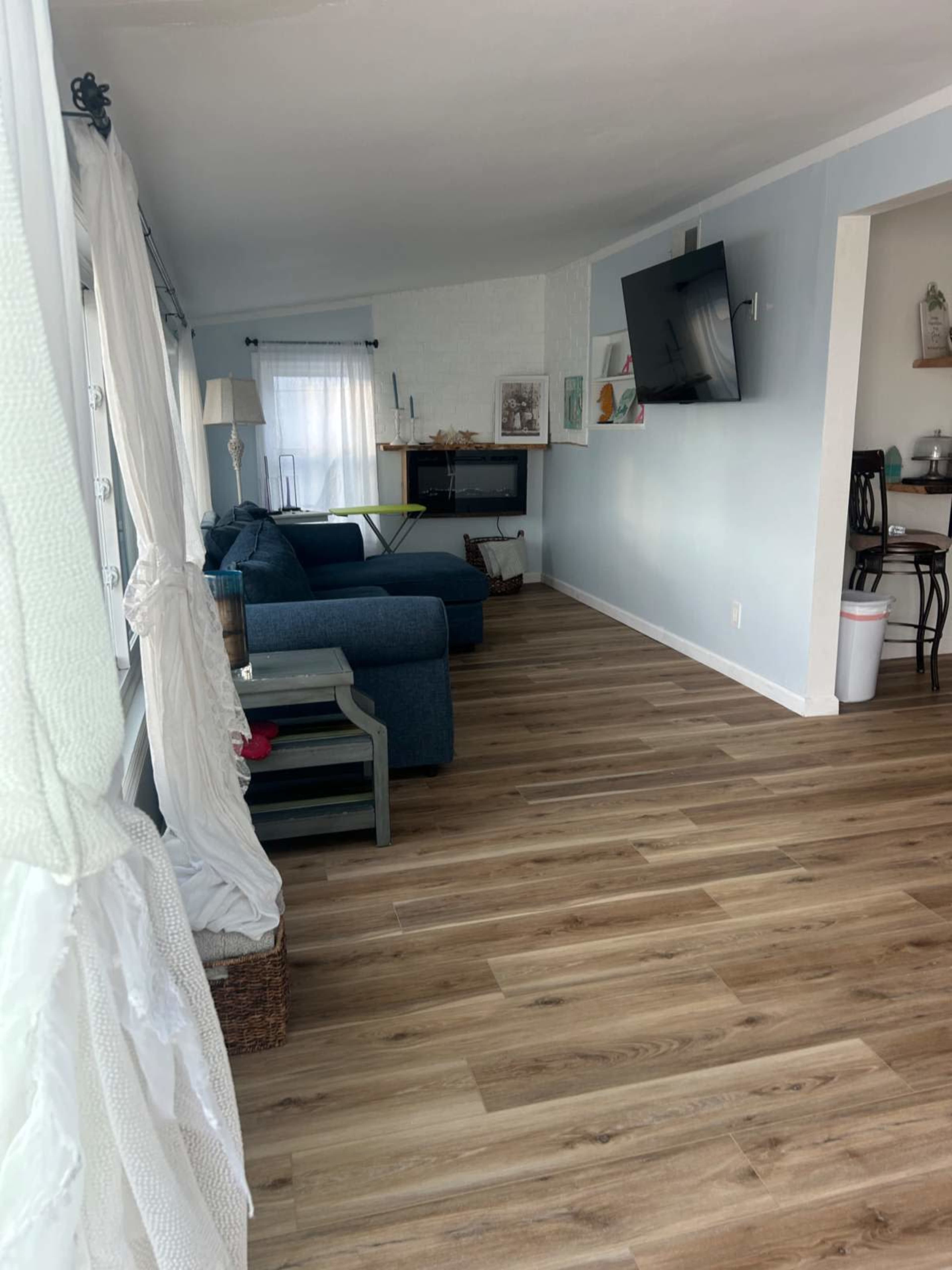 The image shows a living room with blue sofas, light wood flooring, and a mounted television on the wall.