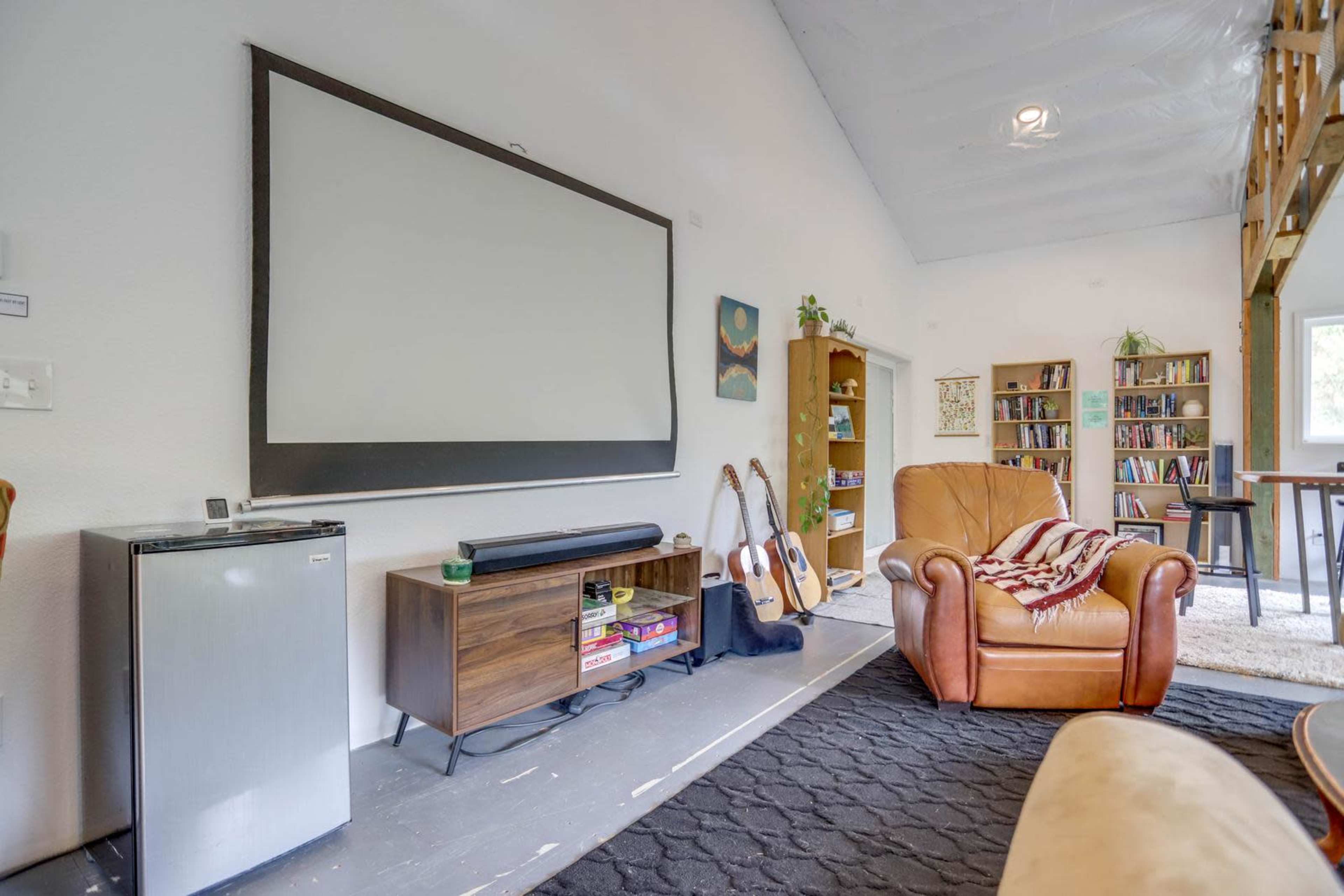 A living area features a large projection screen, a small refrigerator, a wooden coffee table, a guitar, and a brown leather armchair beside a bookshelf filled with books.
