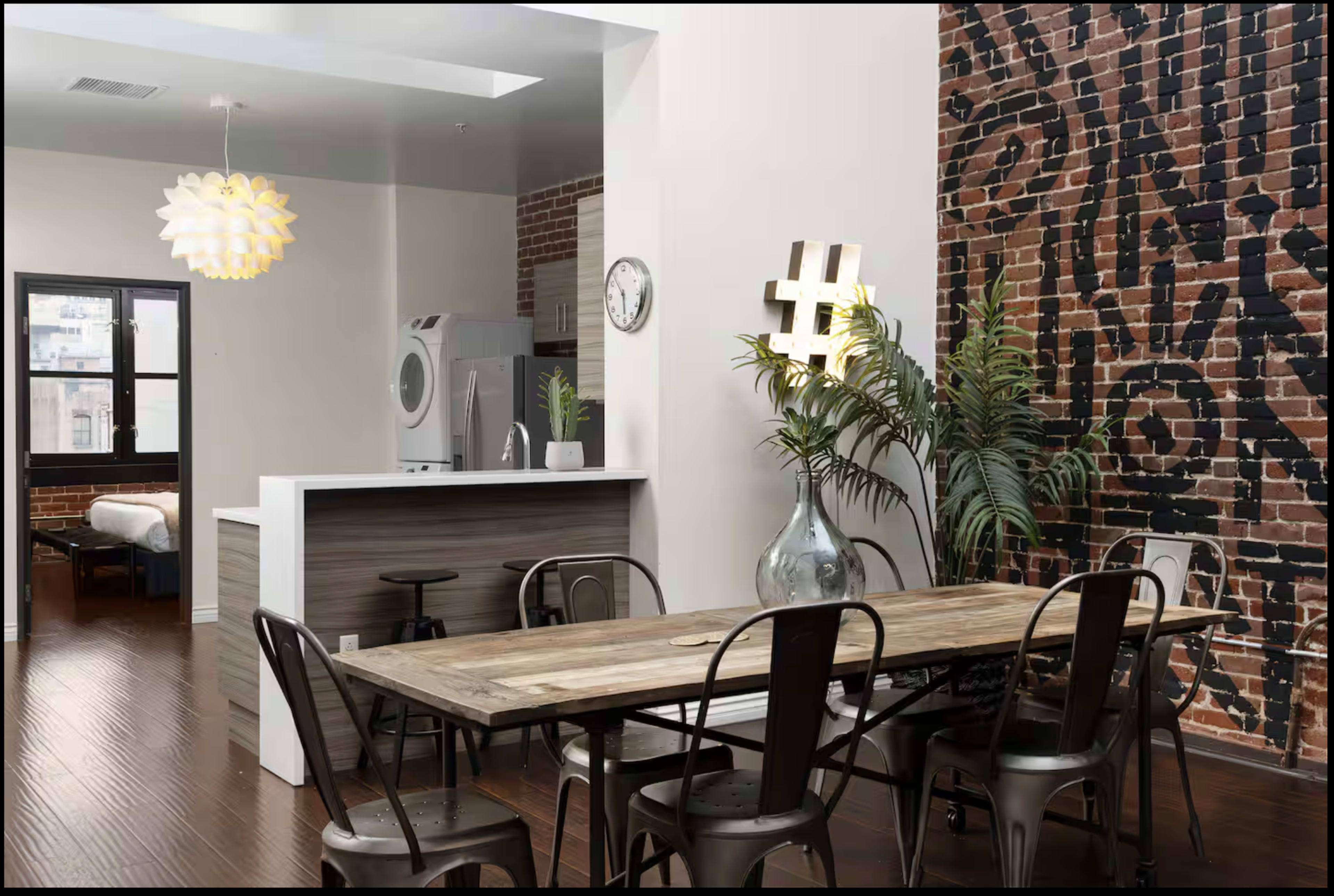 The image shows a modern dining area with a wooden table and metal chairs, situated adjacent to a kitchen featuring exposed brick walls and a decorative wall element.