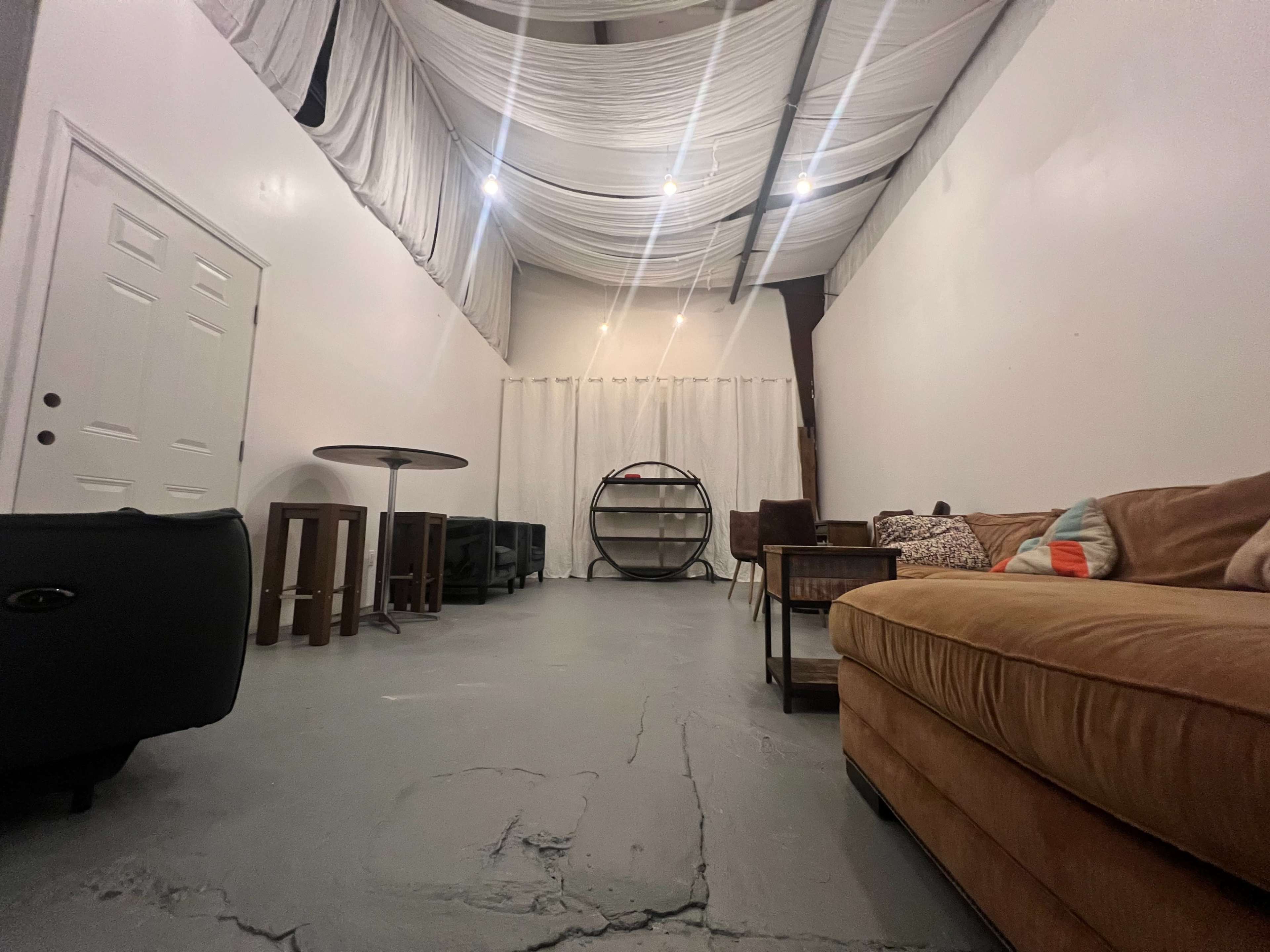 A spacious room features a brown sofa, various seating options, a circular shelf, and white drapery overhead.