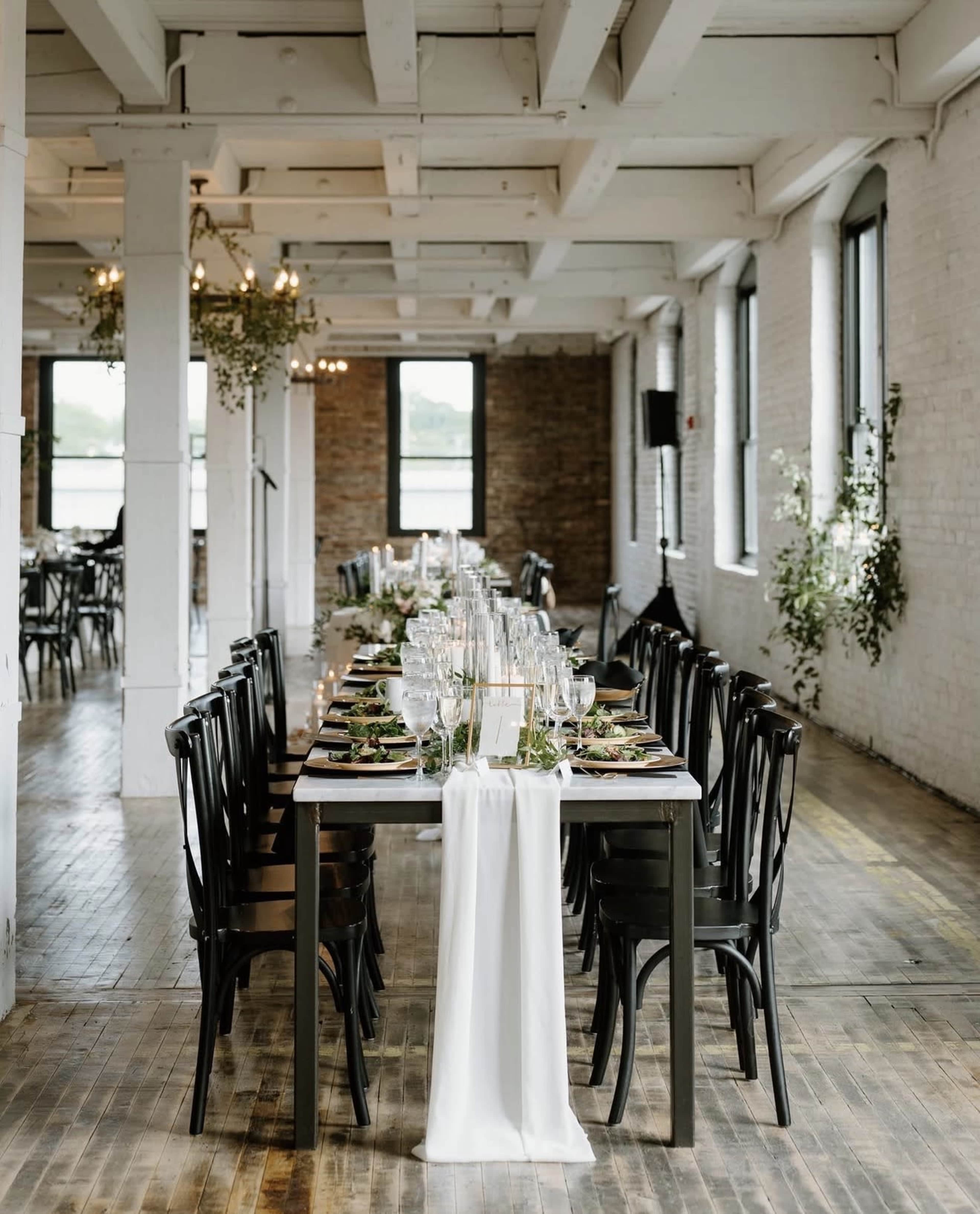 A long dining table is elegantly set with plates, glasses, and decorative greenery in a spacious, brightly lit room with exposed brick walls and large windows.