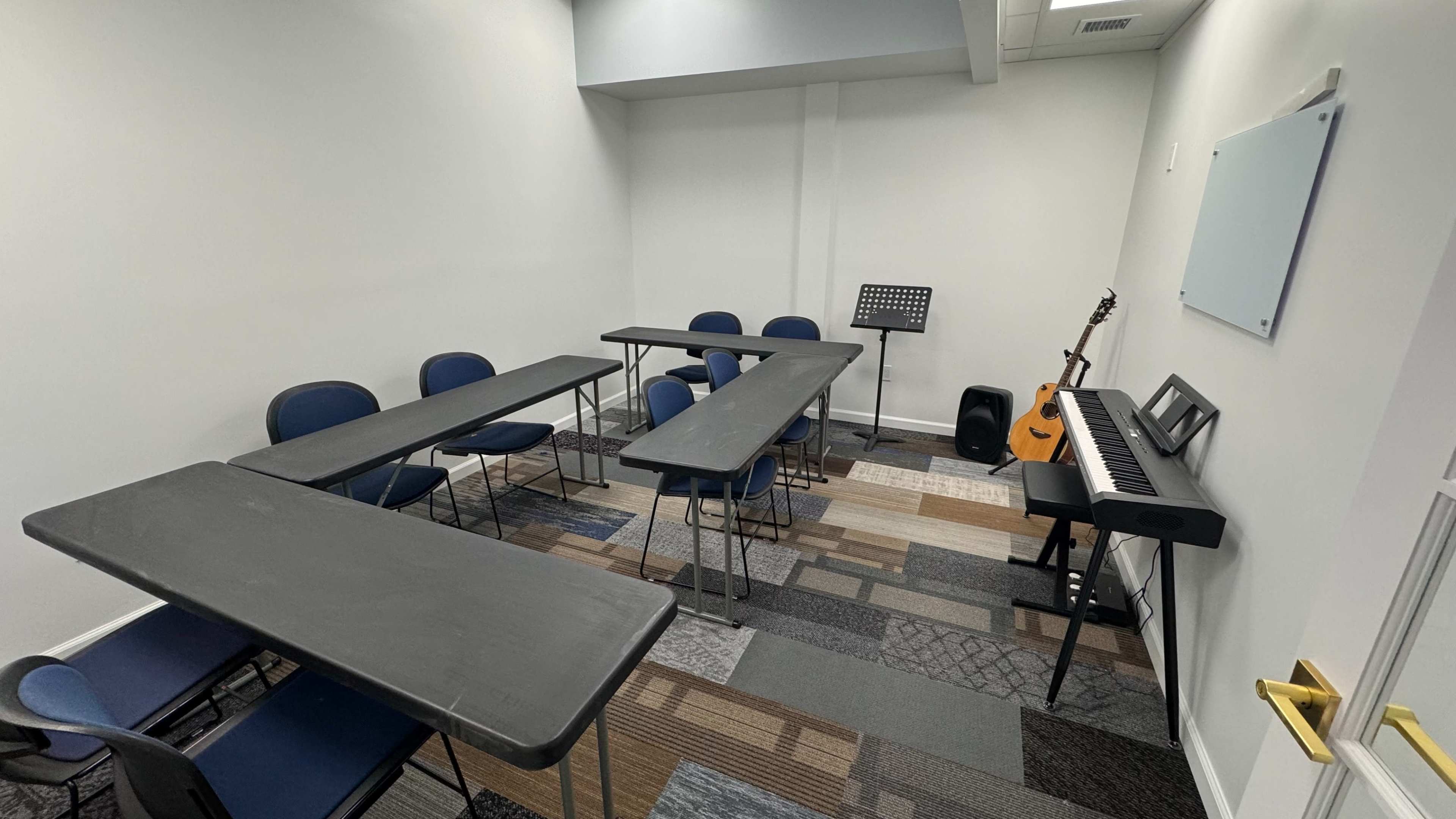A small music room with several tables arranged in a U-shape, blue chairs, a music stand, an acoustic guitar, and a keyboard against the wall.