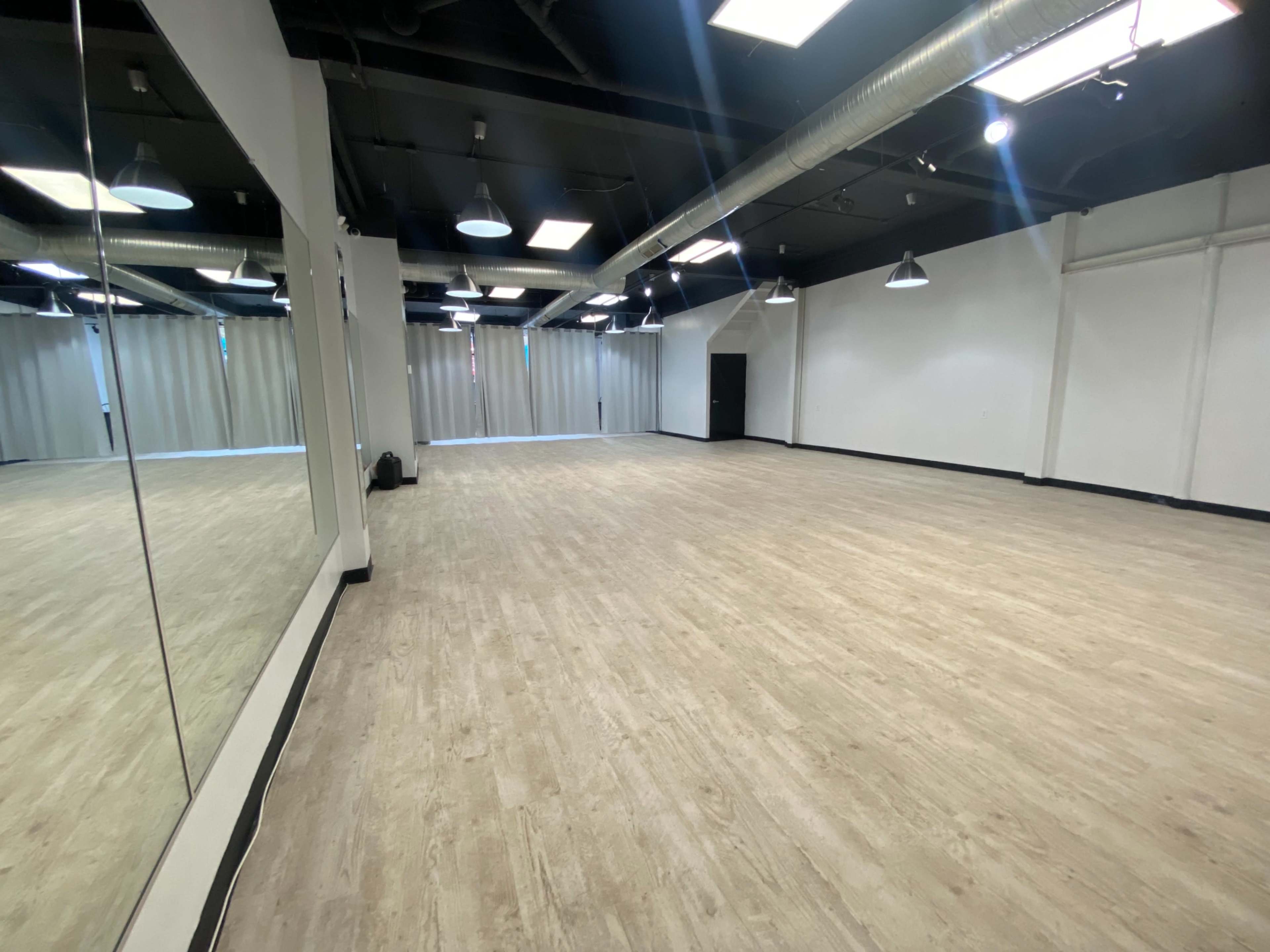 A spacious, empty studio with wooden flooring and large mirrors on one wall, illuminated by overhead lighting.