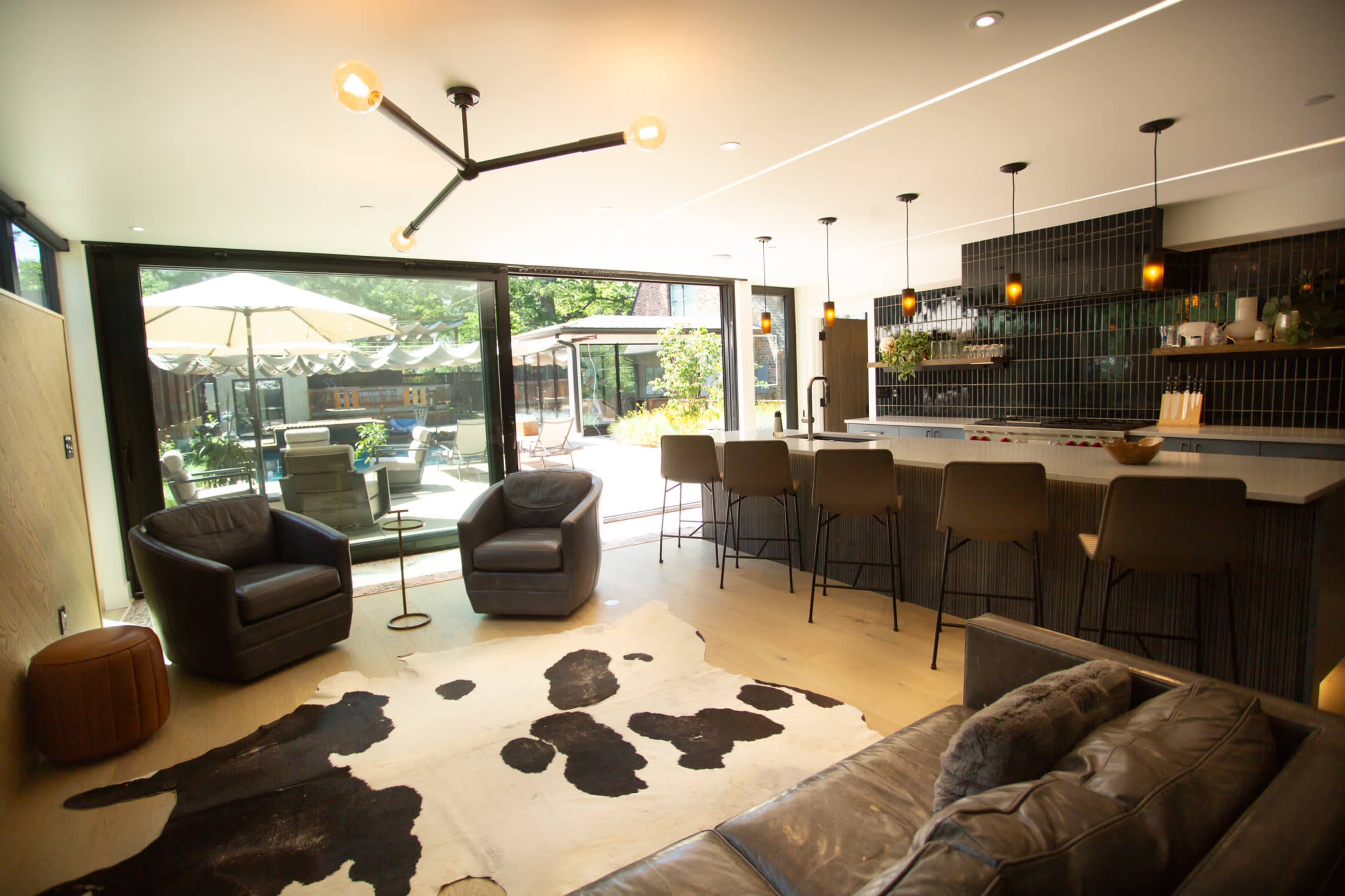 A modern interior features a bar area with stools, a seating area with leather chairs, and large glass doors opening to an outdoor space with umbrellas.