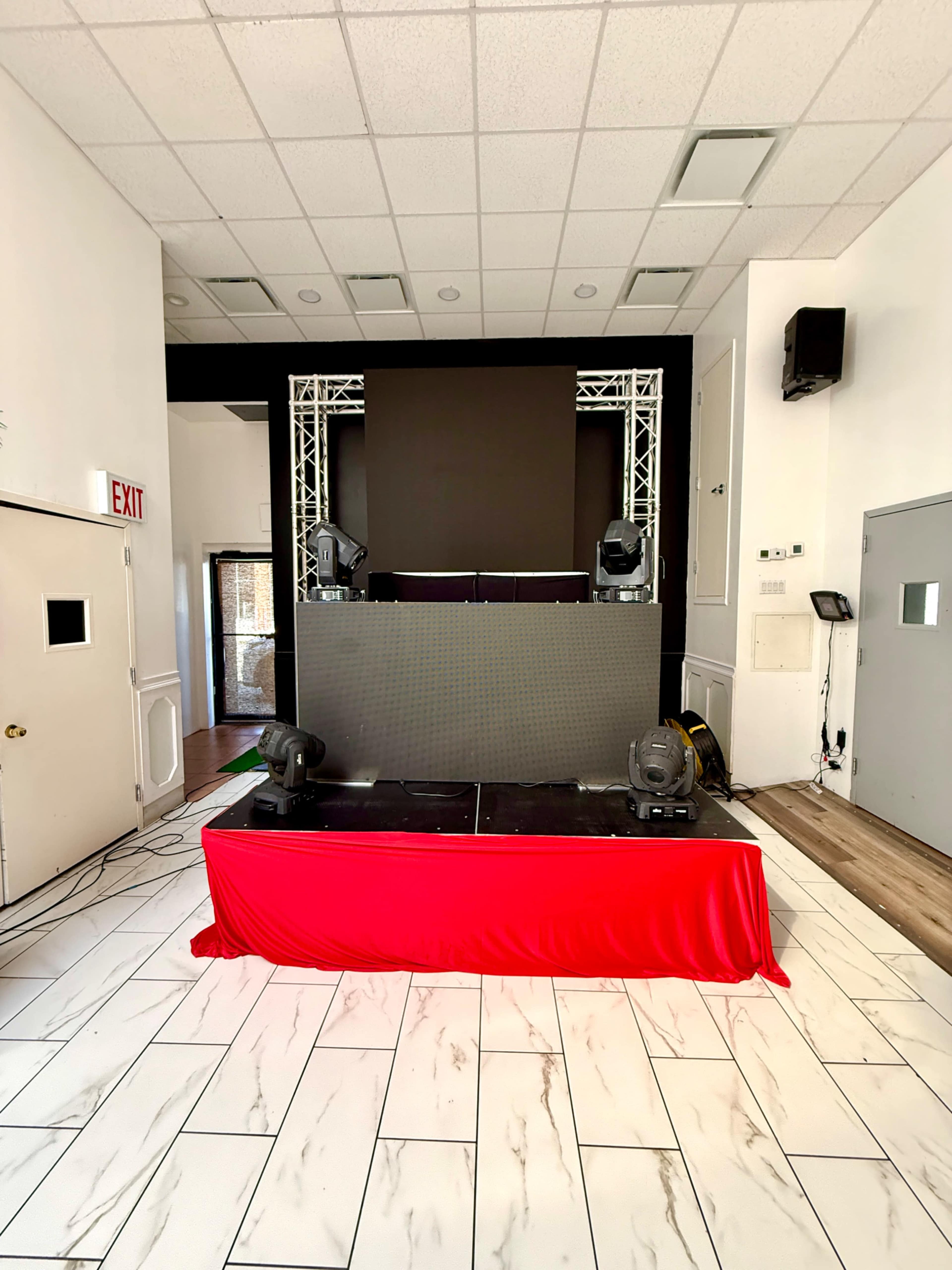 The image shows a performance stage set up with a large screen, projector lights, and a red tablecloth in a room featuring white walls and a tiled floor.