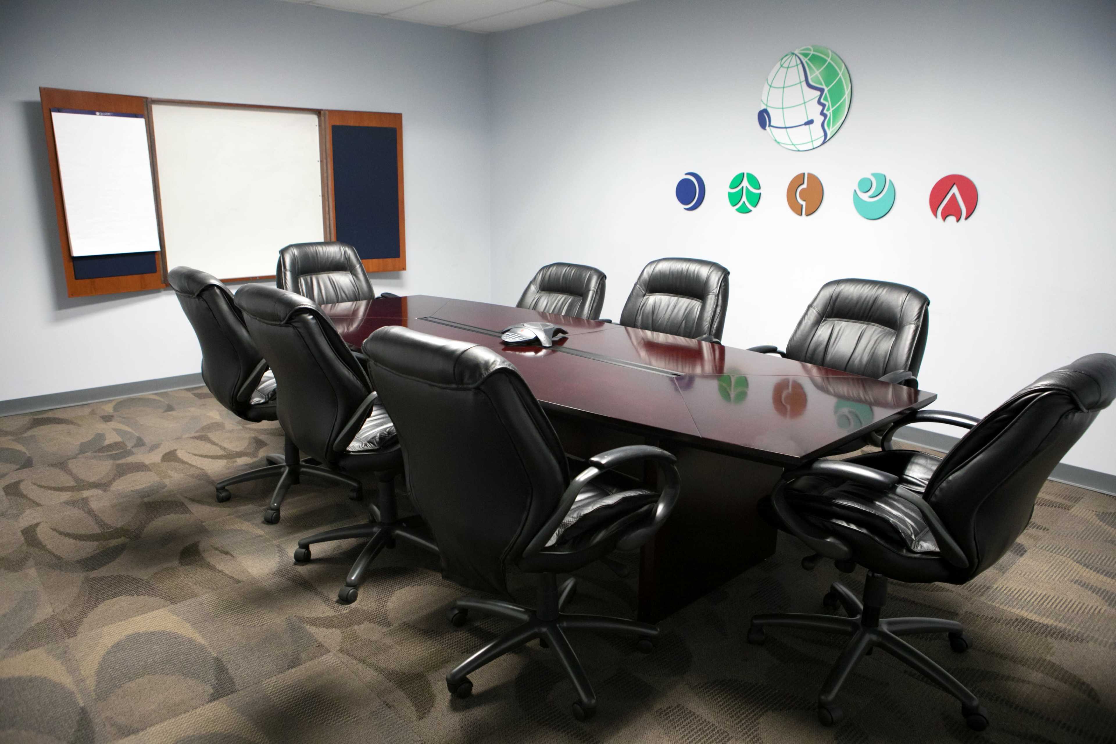 A conference room features a long wooden table surrounded by black leather chairs, with wall decorations of various logos.