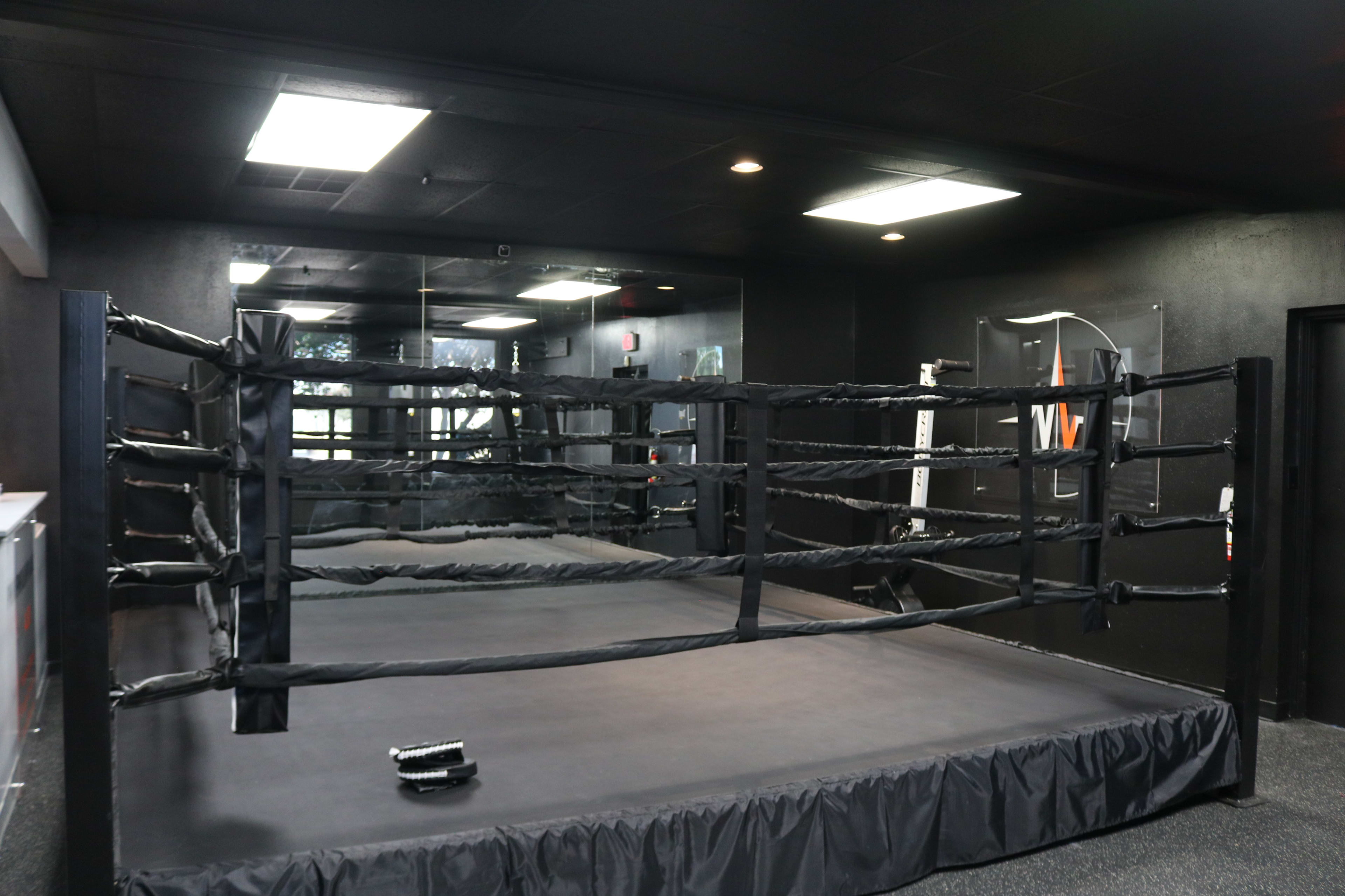 An empty boxing ring set up in a dimly lit gym with black walls and a mirrored backdrop.