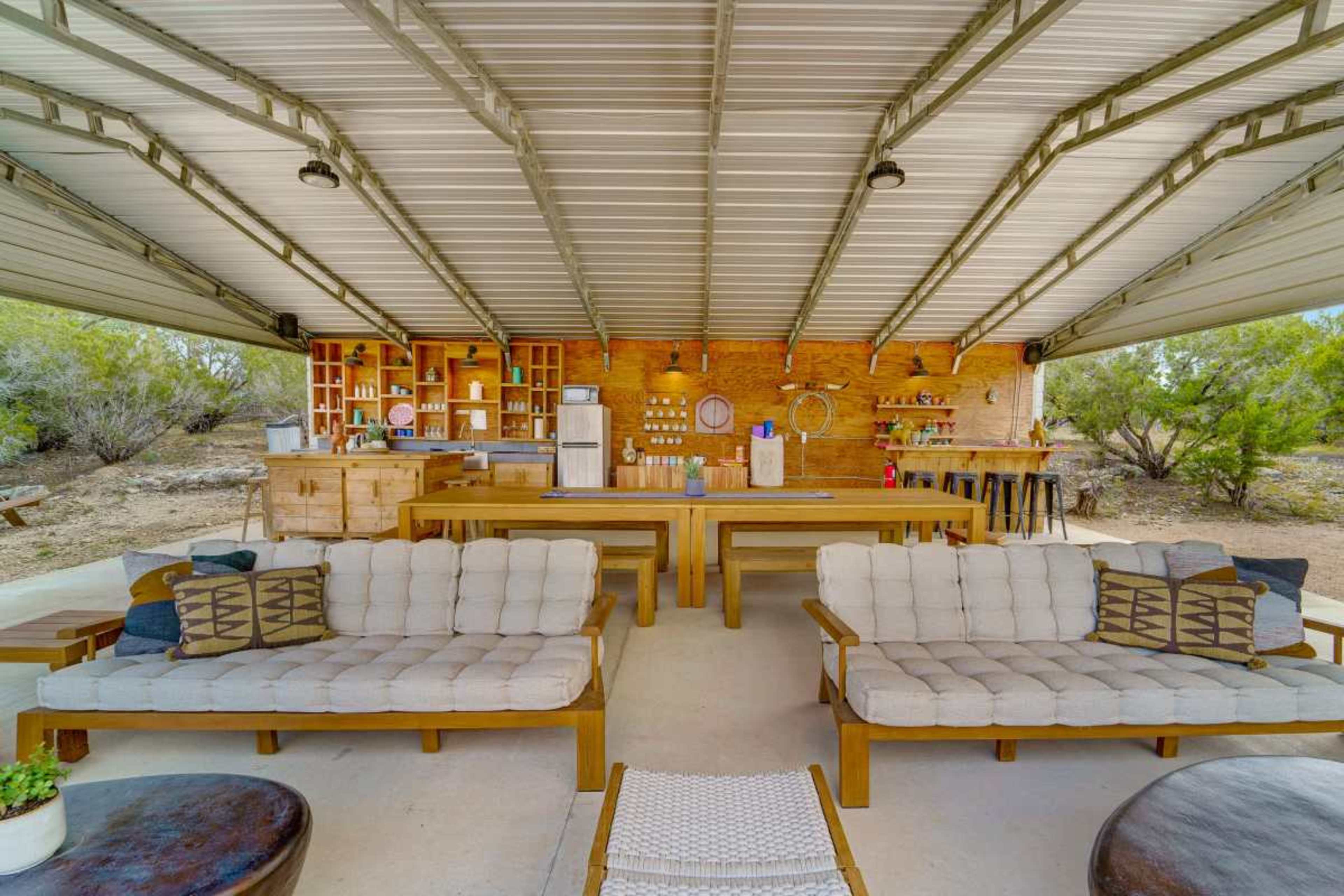 The image shows a spacious outdoor lounge area with wooden furniture, a bar counter in the background, and a covered roof.