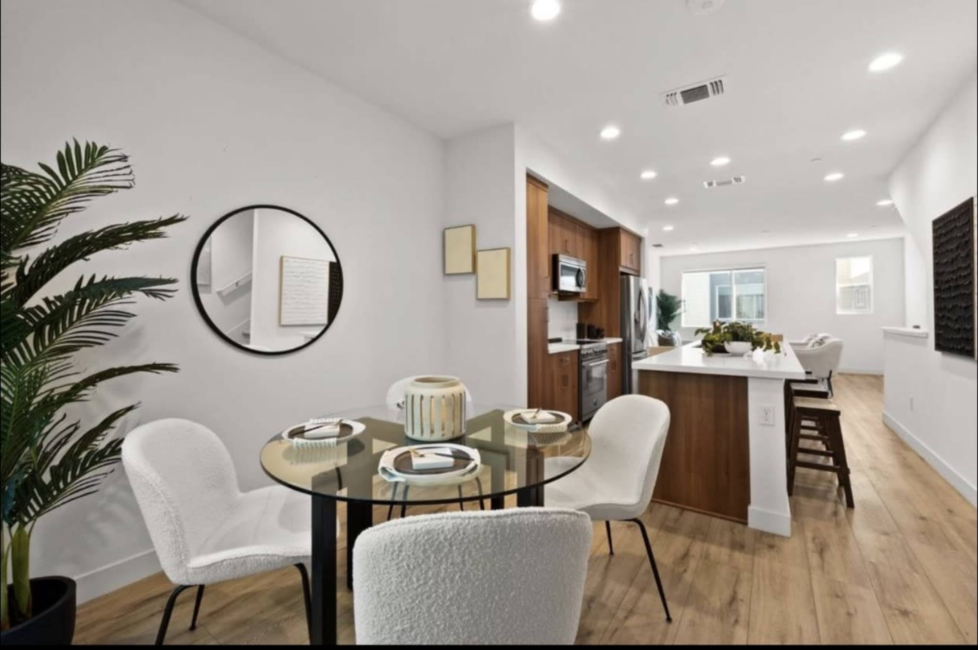 A dining area features a round glass table surrounded by white upholstered chairs, with a mirror and potted plant nearby, adjacent to a kitchen with wooden cabinetry and modern appliances.