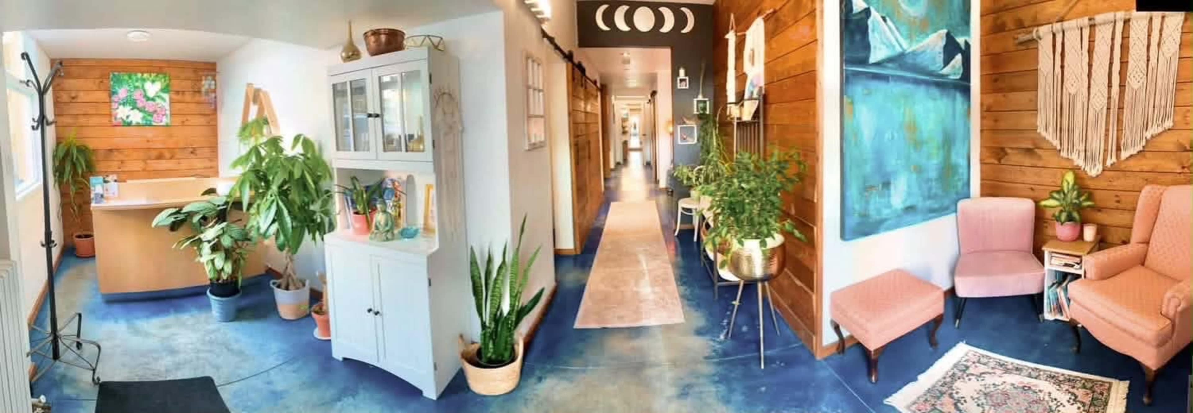 The image shows a bright interior with a reception area featuring plants, a hallway with artwork, and a cozy seating nook with pink chairs.