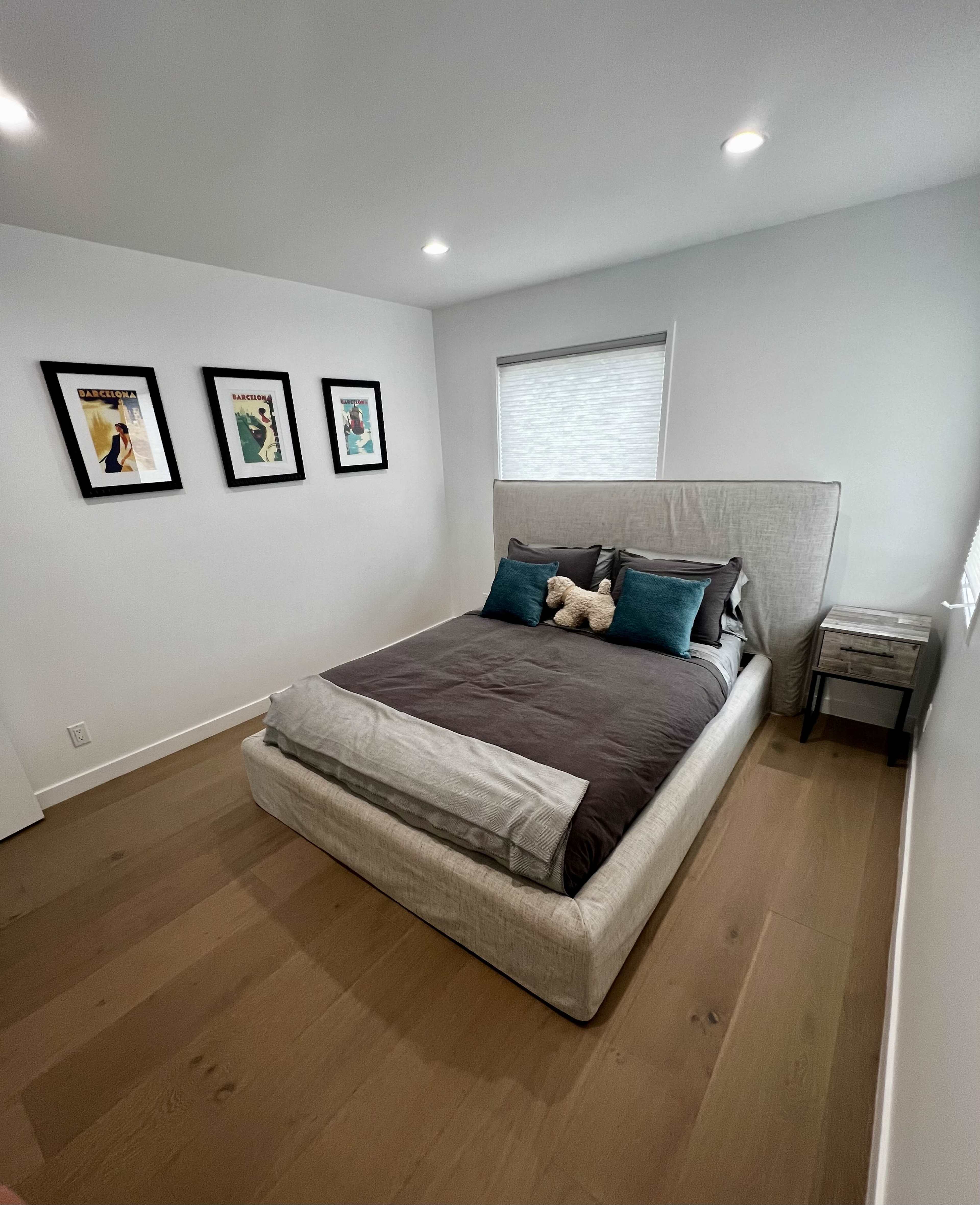 A simple bedroom features a bed with a light-colored headboard, dark bedding, and teal accent pillows, complemented by framed artwork on the walls.
