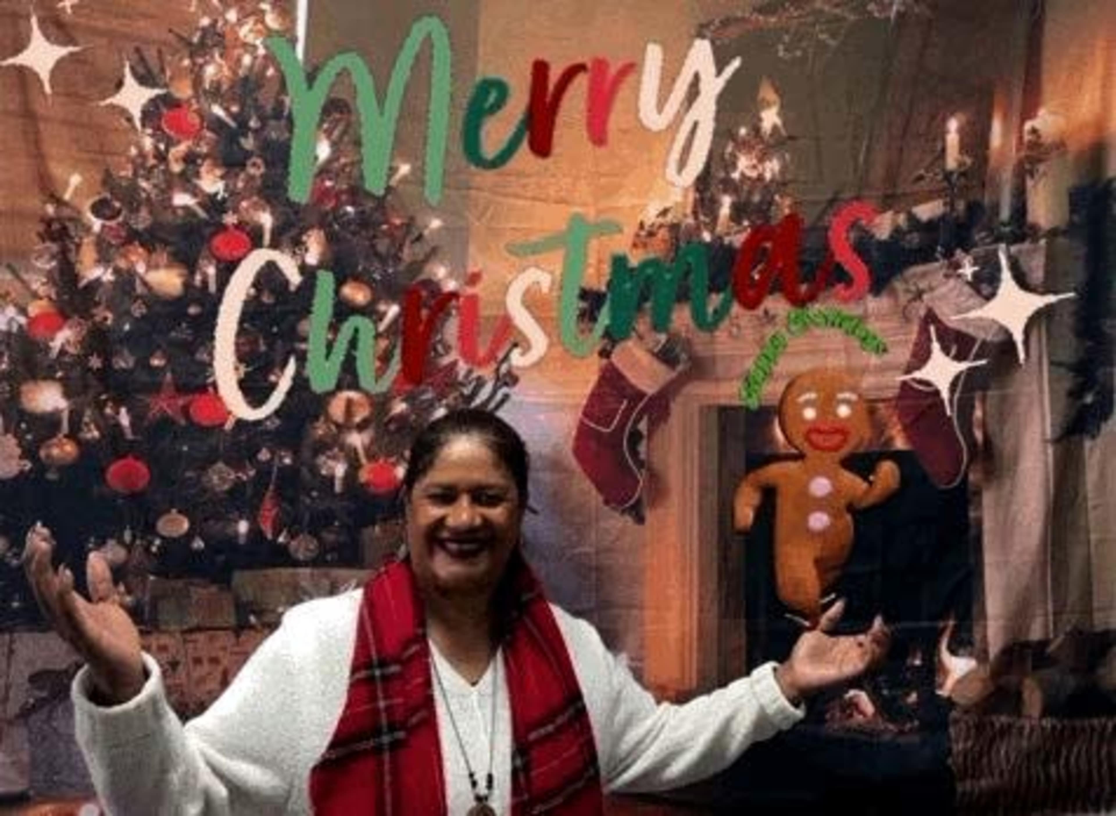A person stands with arms outstretched in front of a decorated Christmas tree and a festive backdrop that reads "Merry Christmas."
