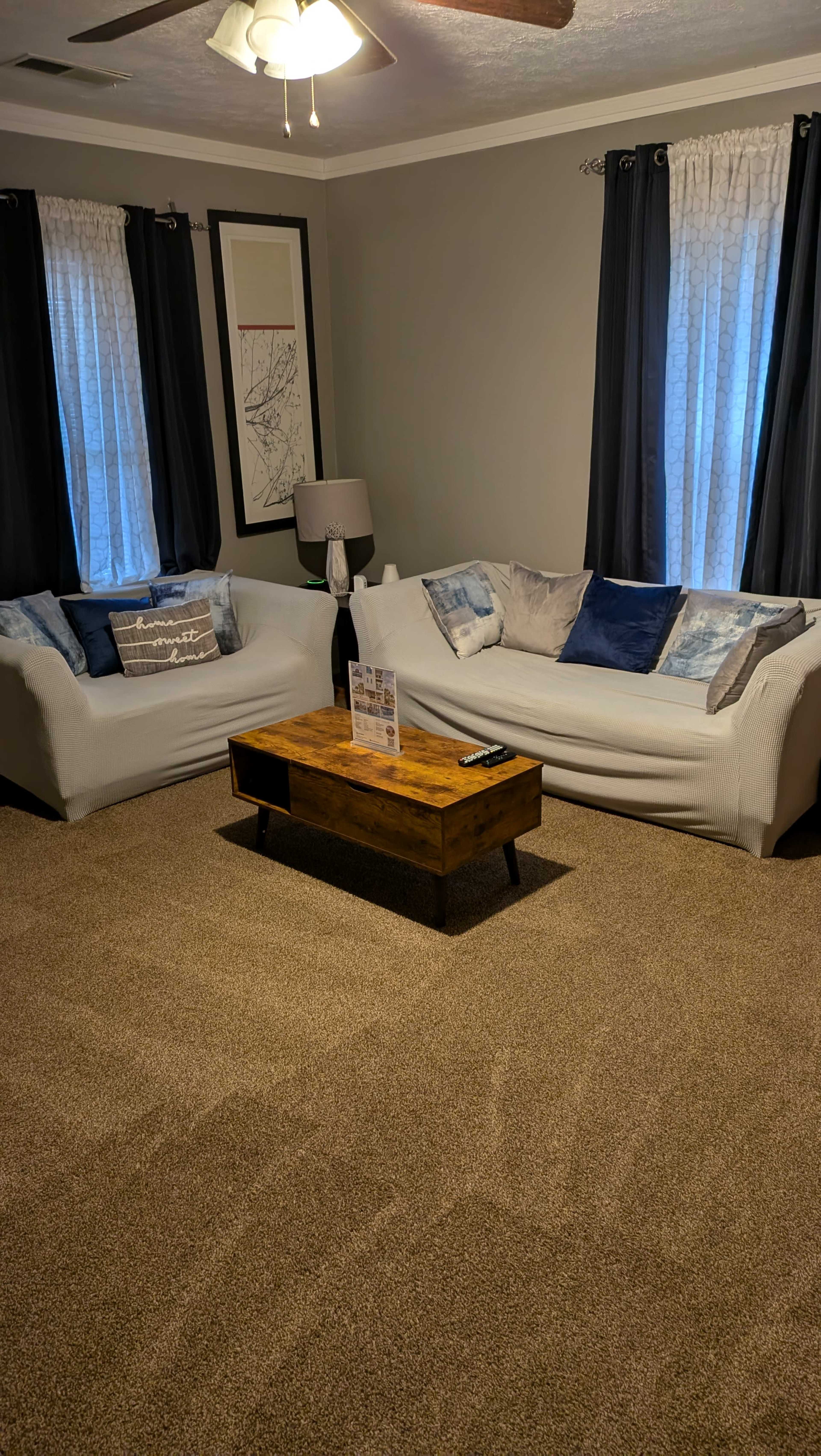 The image shows a living room with two light-colored sofas facing a wooden coffee table, surrounded by curtains and a ceiling fan overhead.