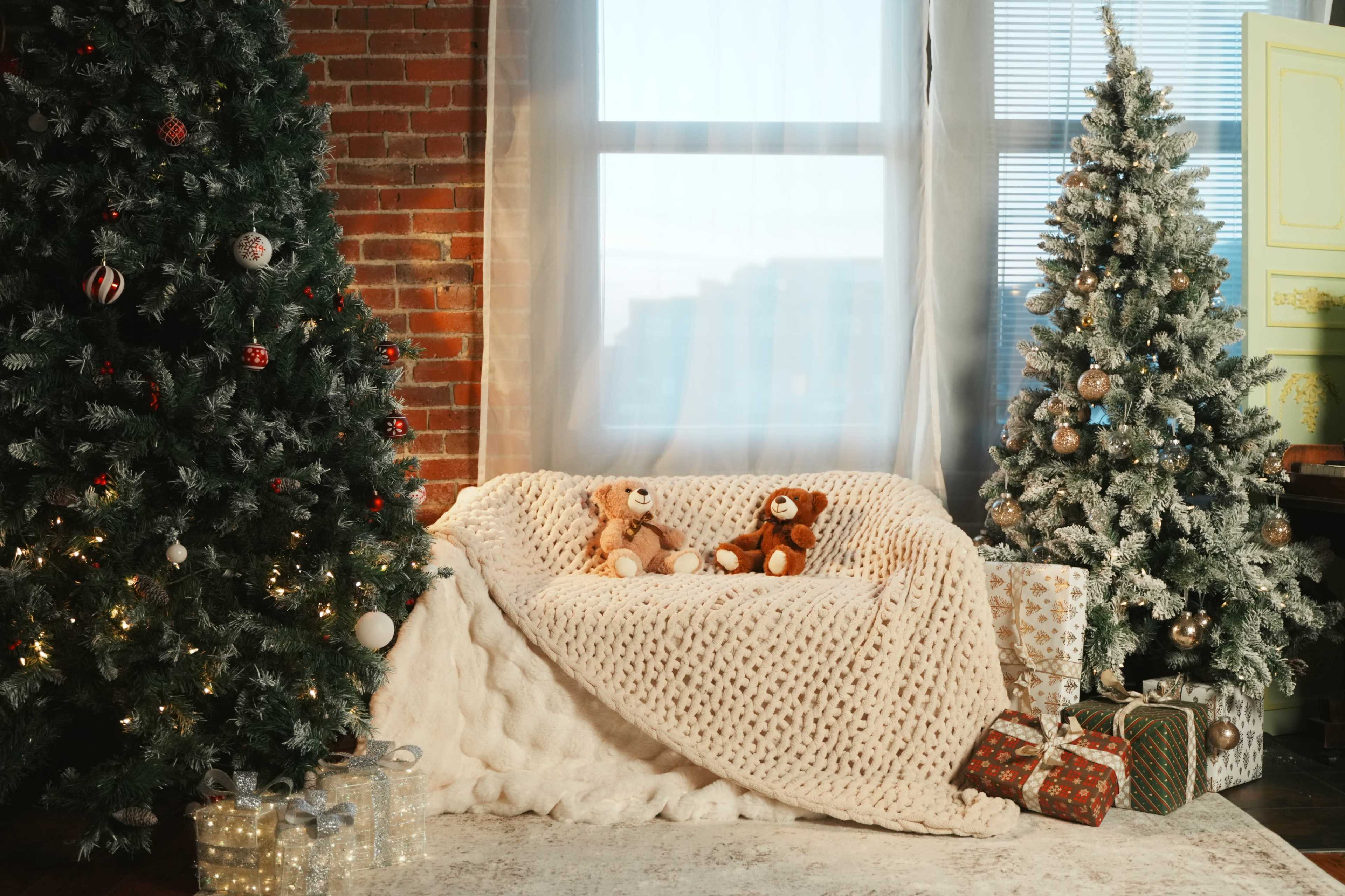 A cozy living room scene features a knitted blanket on a sofa surrounded by Christmas trees, stuffed animals, and wrapped gifts.