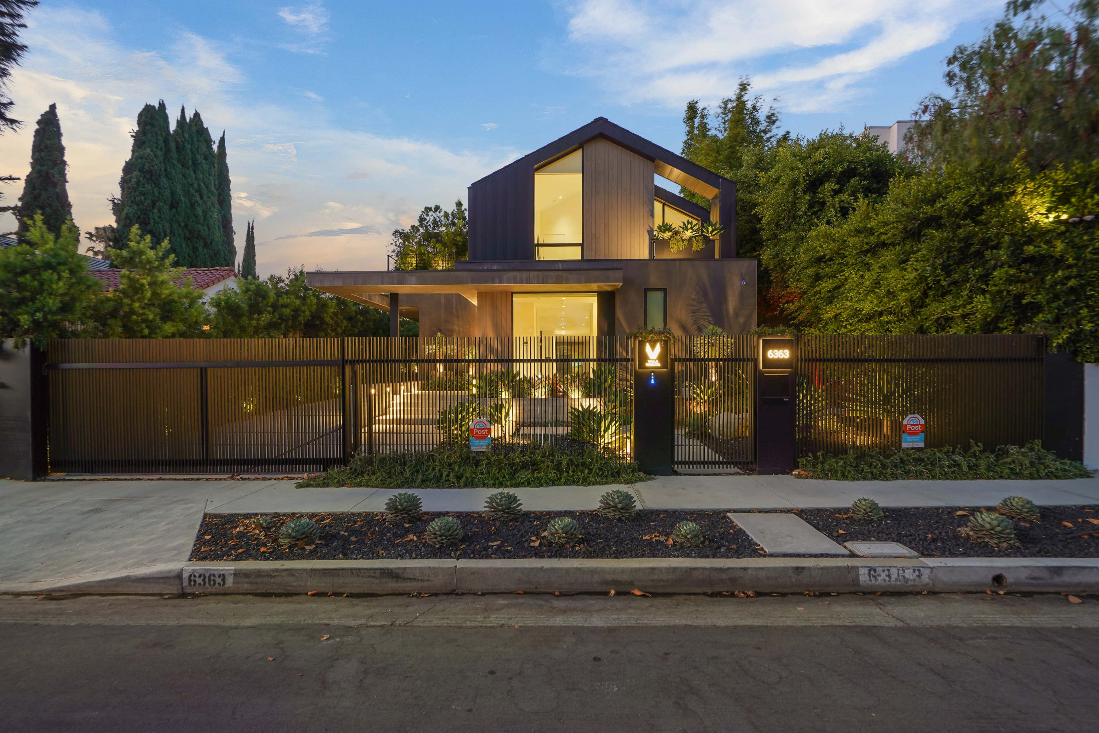 A modern two-story house with a geometrically angled roof and large windows is set behind a black metal fence, surrounded by greenery and a landscaped yard.