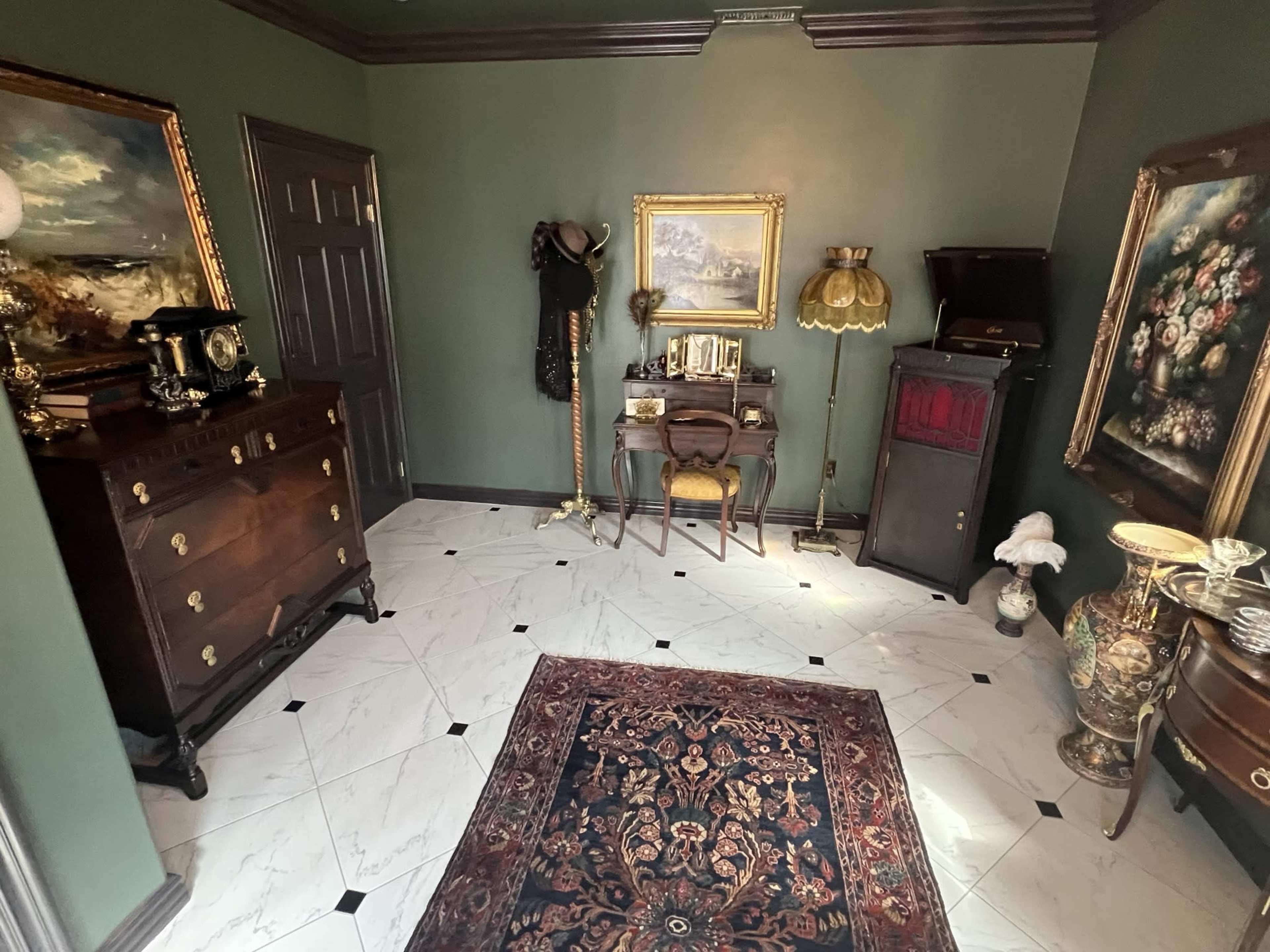 The image shows a vintage-style entryway featuring dark green walls, a marble floor, and various antique furniture pieces, including a dresser, a small table with a mirror, and a record player.