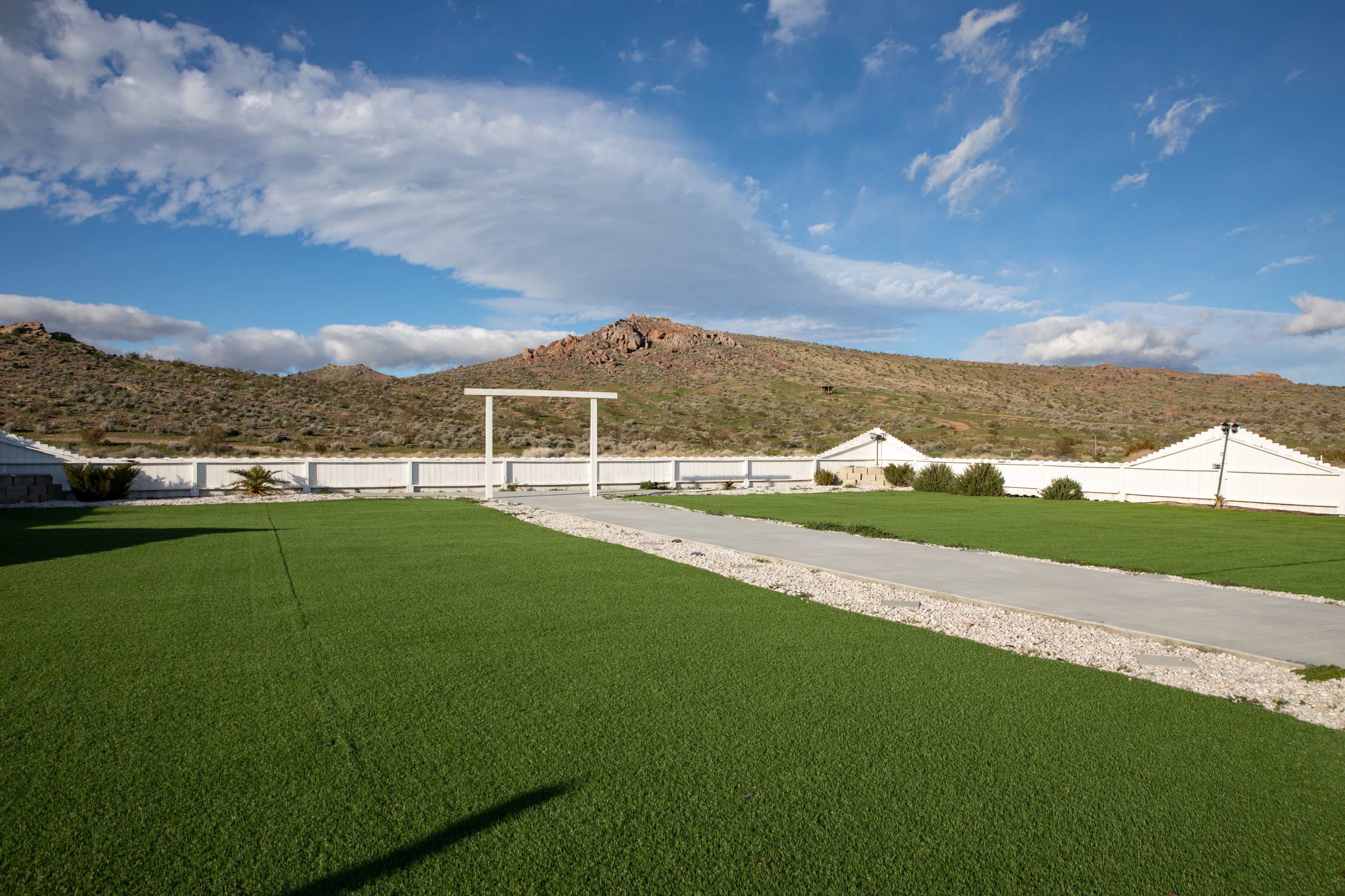 A large expanse of green grass is shown in the foreground, bordered by a gravel path and surrounded by mountains under a partly cloudy sky.