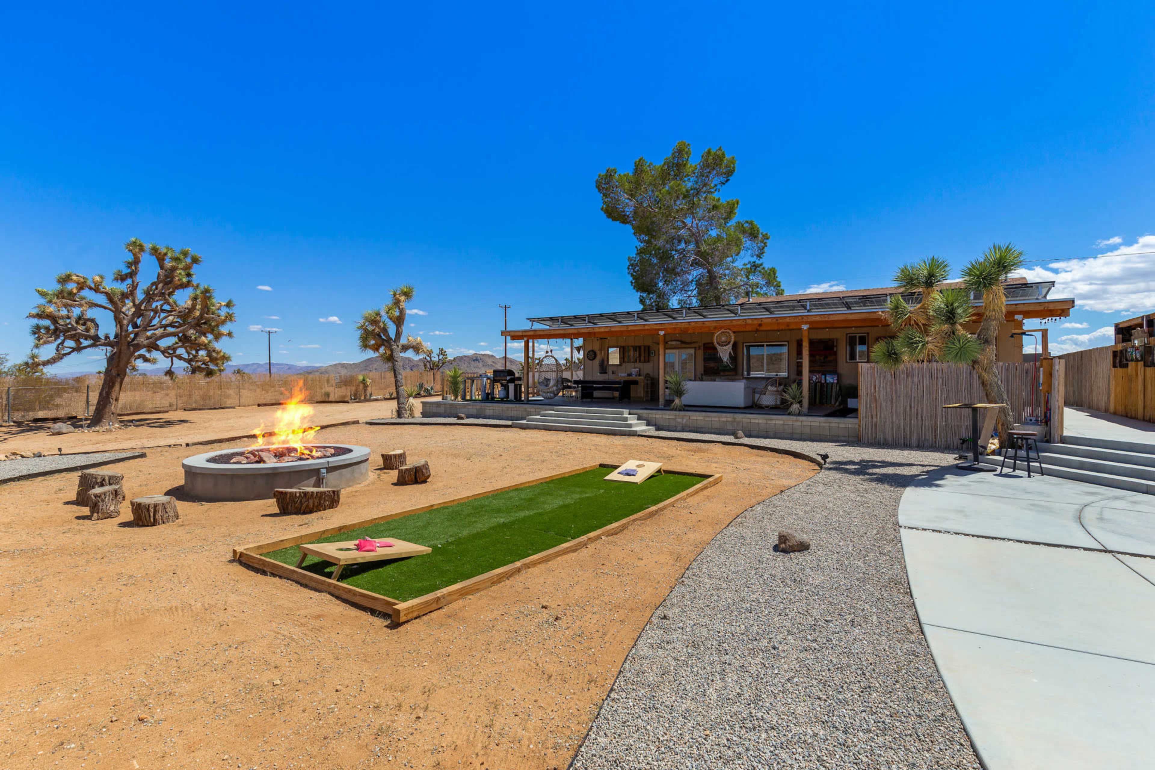 The image shows a desert outdoor space featuring a fire pit, a grassy area for games, and a rustic building surrounded by Joshua trees and a clear blue sky.