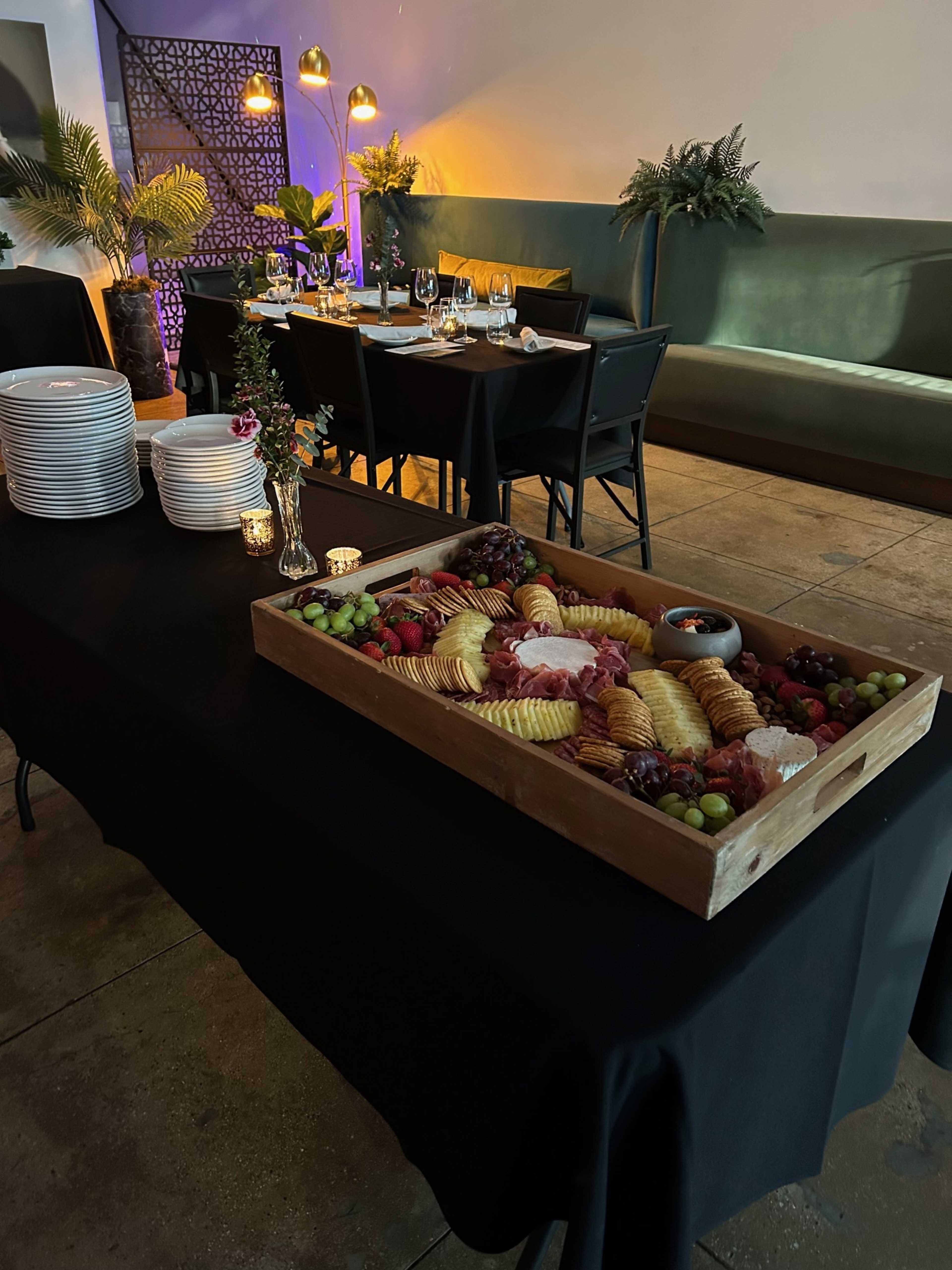 A wooden serving tray filled with an assortment of cheeses, meats, and fruits is placed on a black tablecloth beside a dining setup with tables and chairs in a dimly lit room.