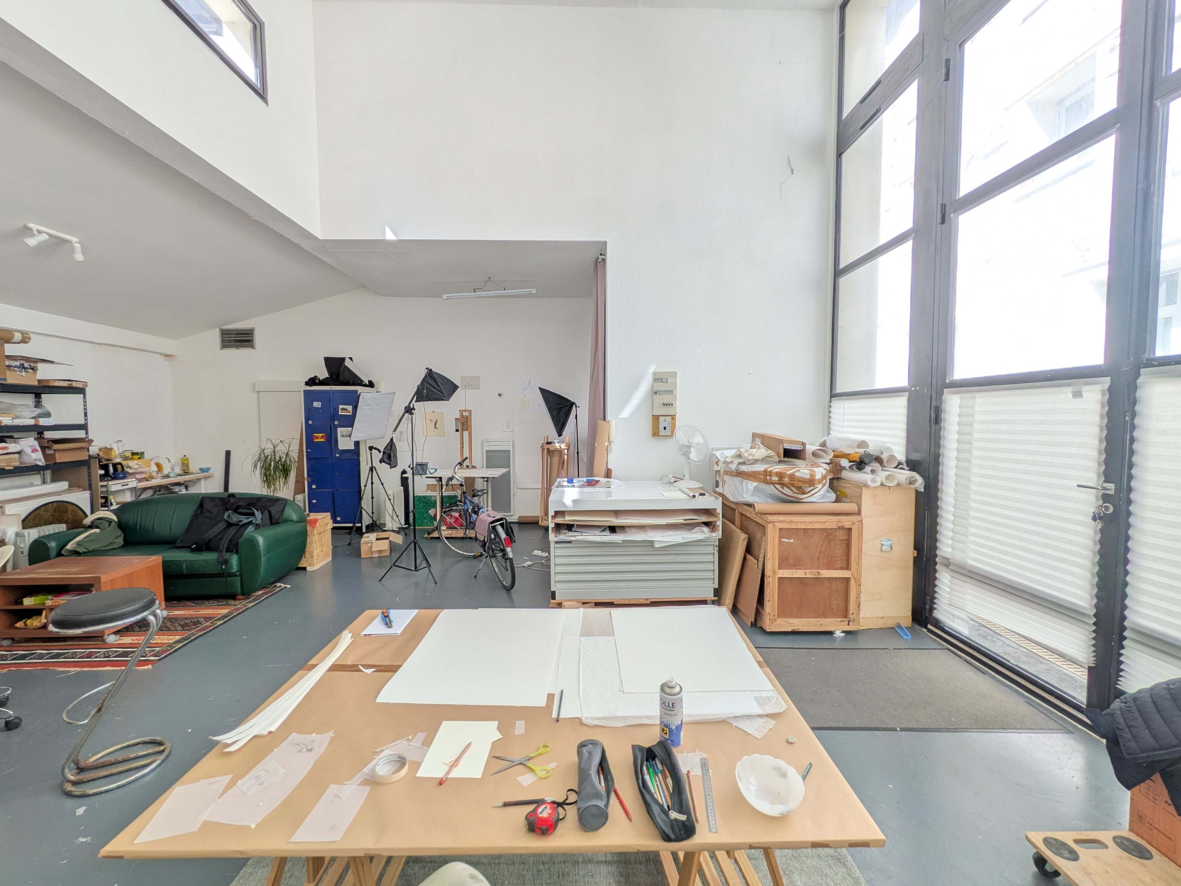 The image shows an artist's studio with a large work table in the foreground, shelves in the background, and natural light coming through large windows.