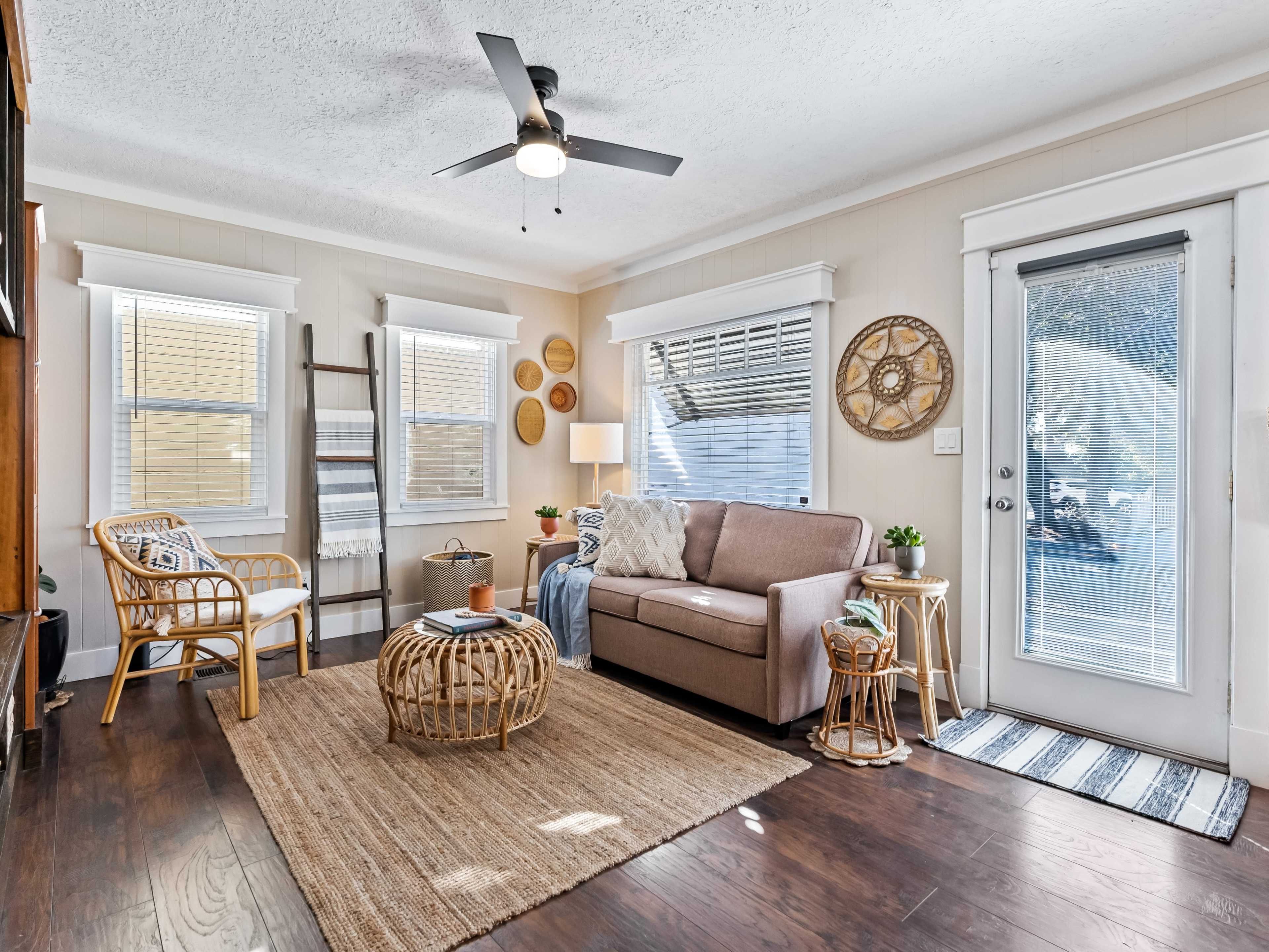 The room features a light-colored living area with a sofa, a round woven coffee table, and large windows allowing natural light to enter.