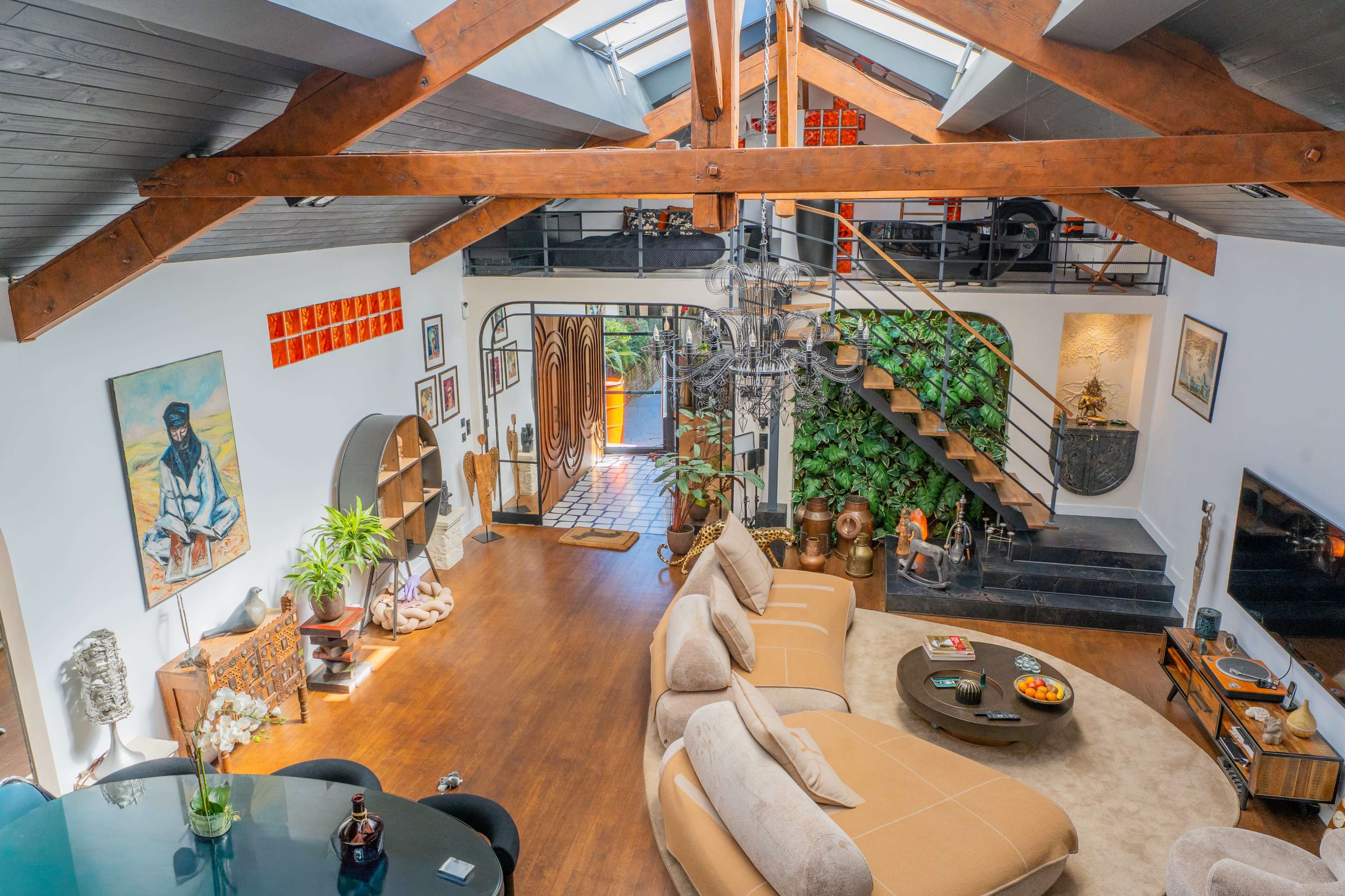 The image shows an open-concept living space with wooden beams, a large sofa, a dining area, and decorative plants, along with artistic elements on the walls and a staircase leading to an upper level.